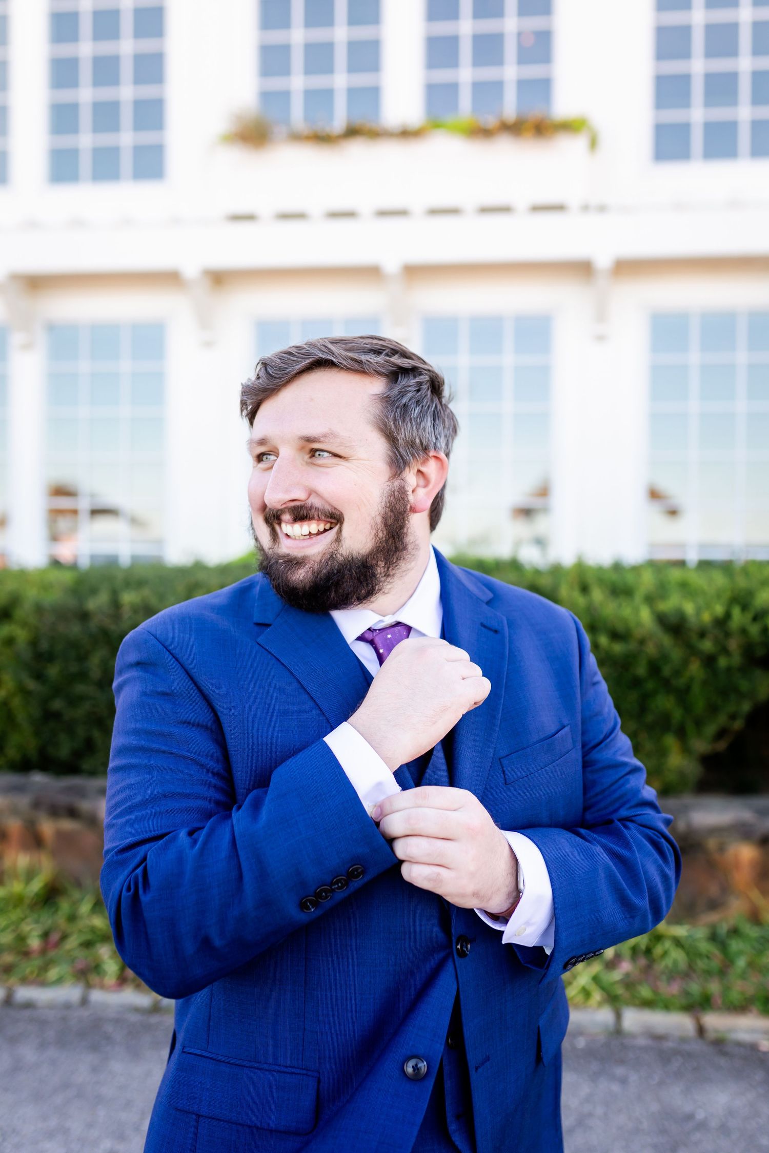 Person in bright blue suit adjusts cuff while smiling outside building.
