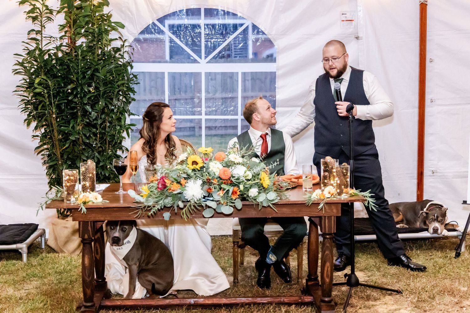 Outdoor wedding reception features farm tables and floral arrangements under a white tent with window panels.