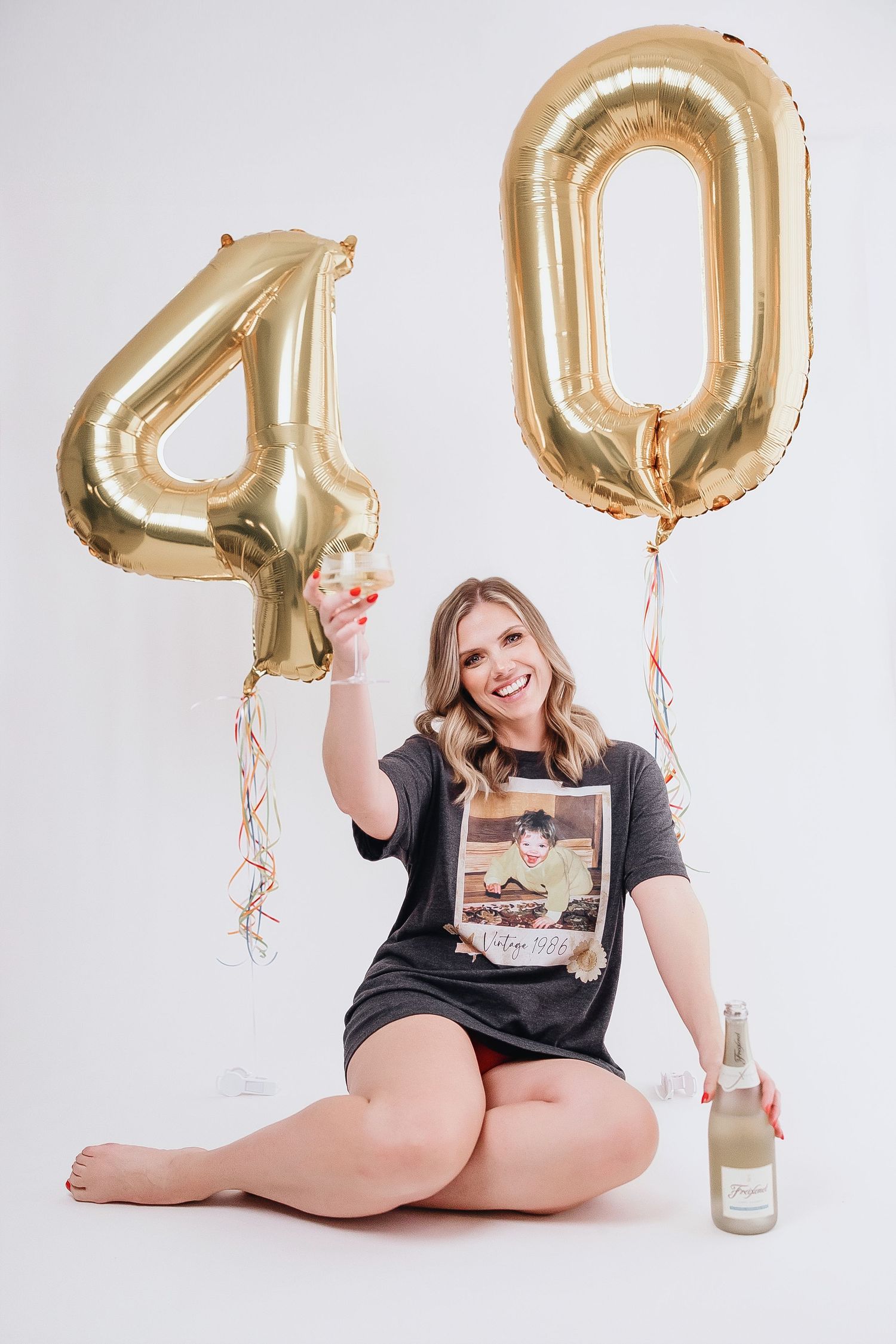 Woman in black t-shirt poses with gold number 40 balloons and champagne bottle against white backdrop.