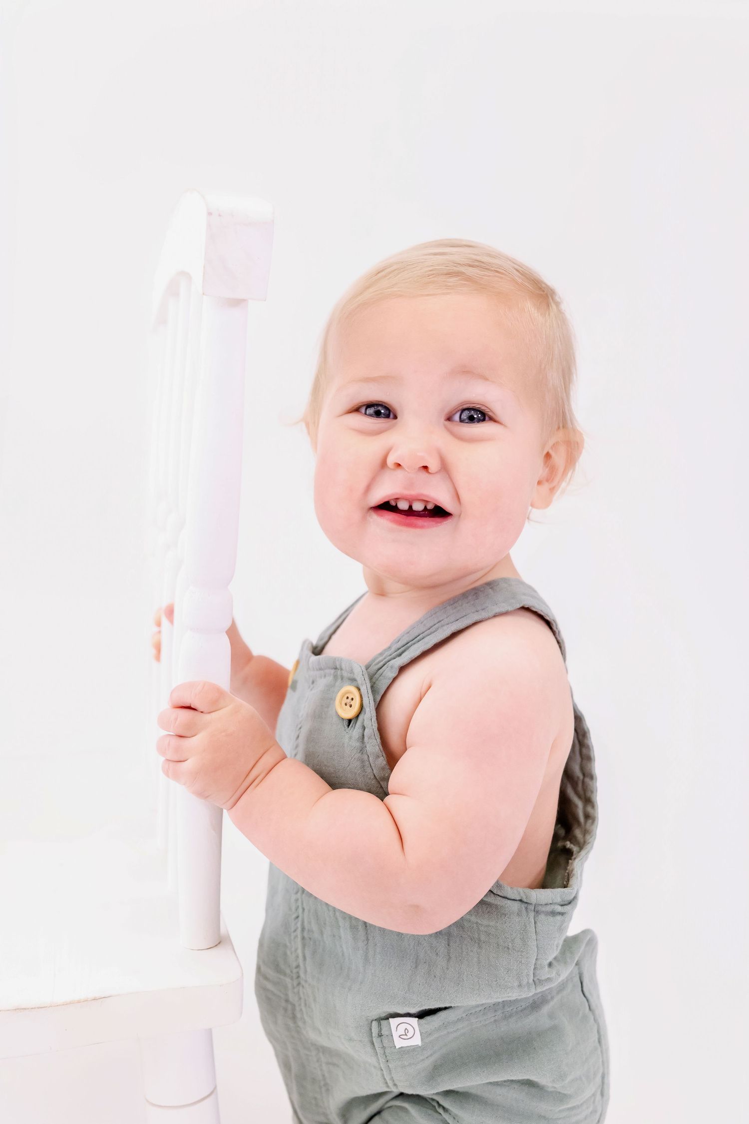 A baby in grey overalls laughs while sitting in a white chair during a photo session.
