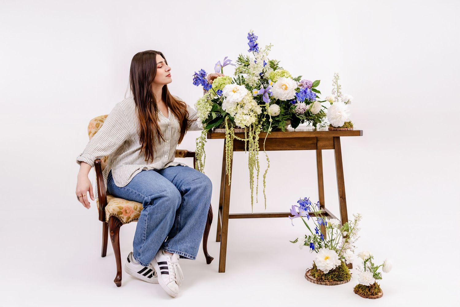 Woman sitting on chair beside wooden table with white and blue floral arrangements on white background.