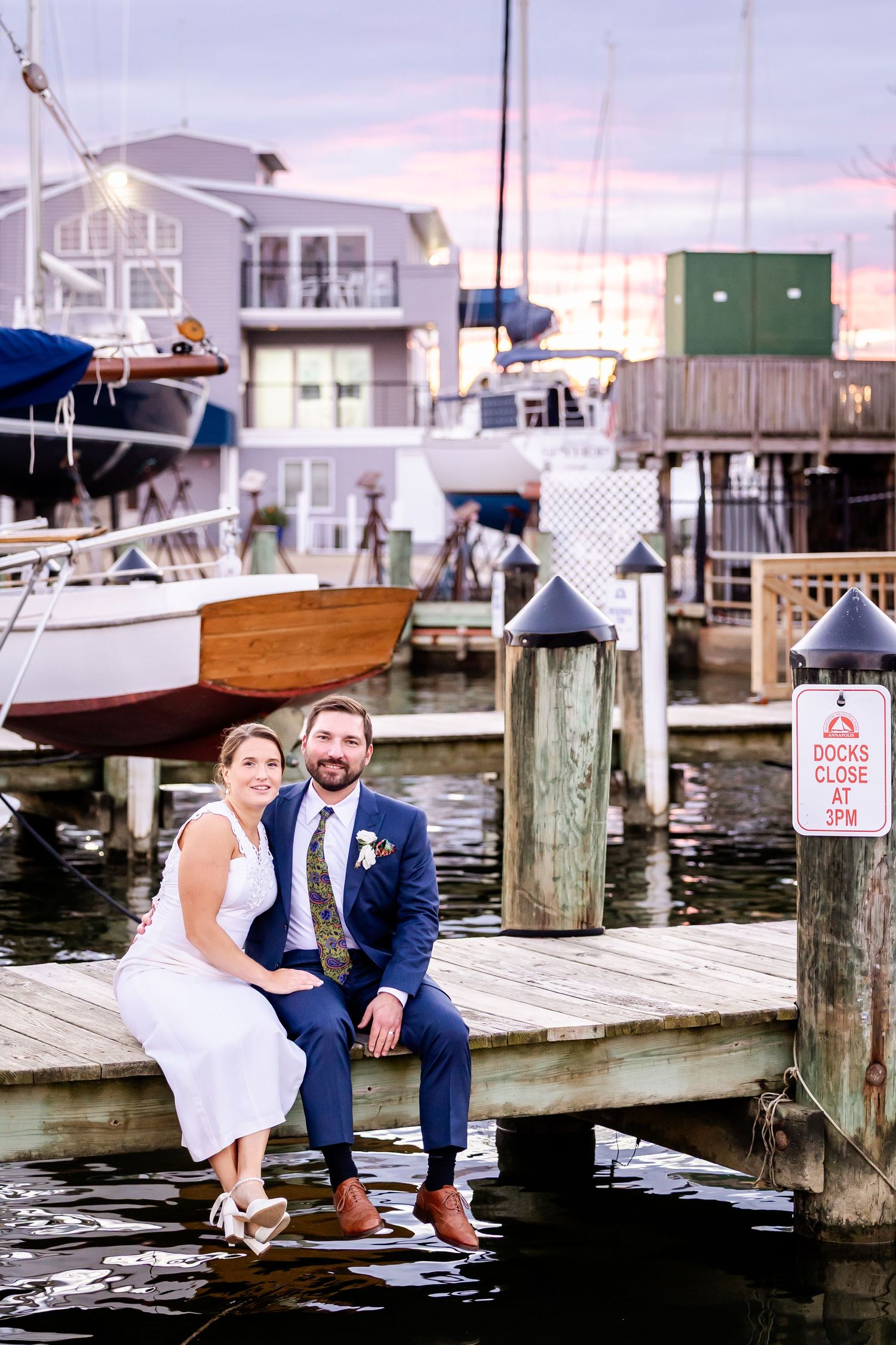 Bride and groom sitting on a marina dock at sunset, with sailboats and waterfront buildings in the background.