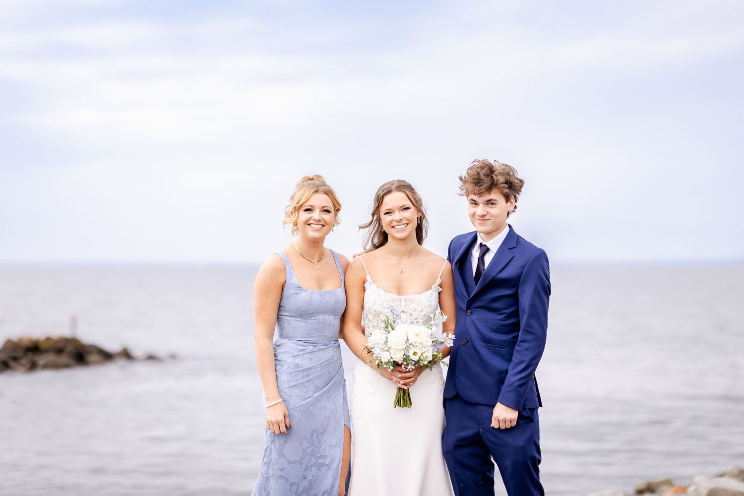 Three people in formal attire stand together by the ocean on an overcast day with a rocky shoreline in the background.