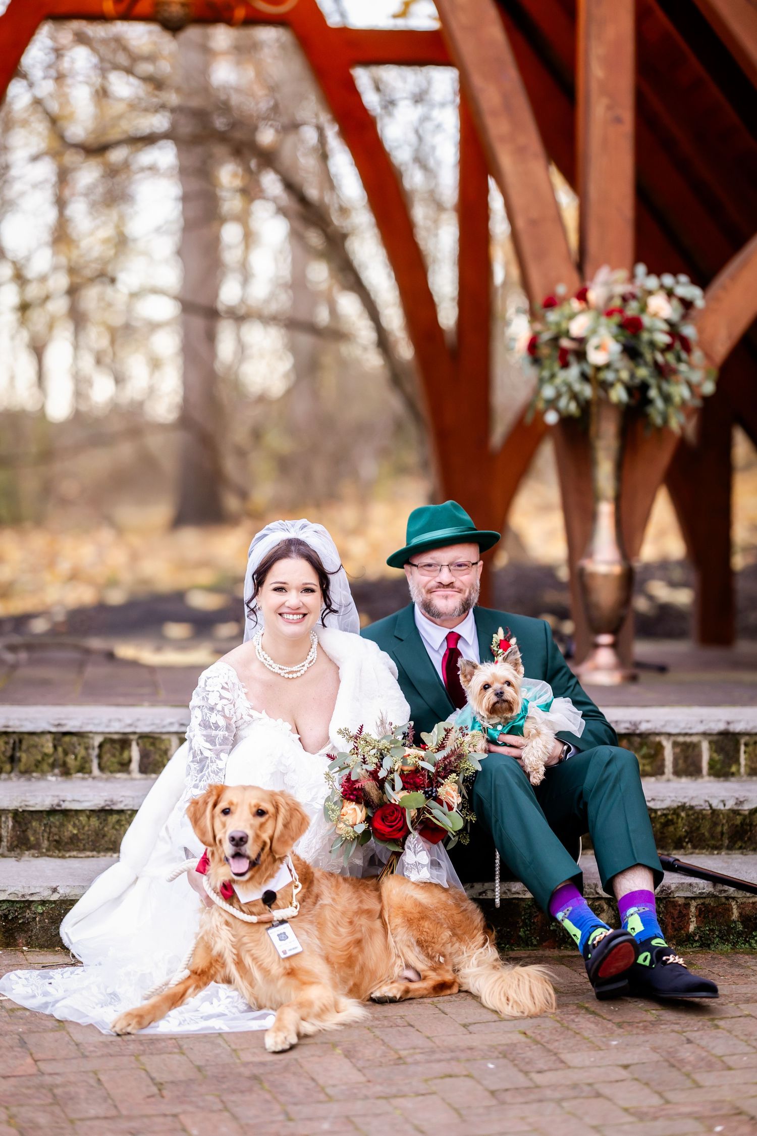 A wedding couple sits with their golden retriever and small dog under a wooden arch decorated with floral arrangements.
