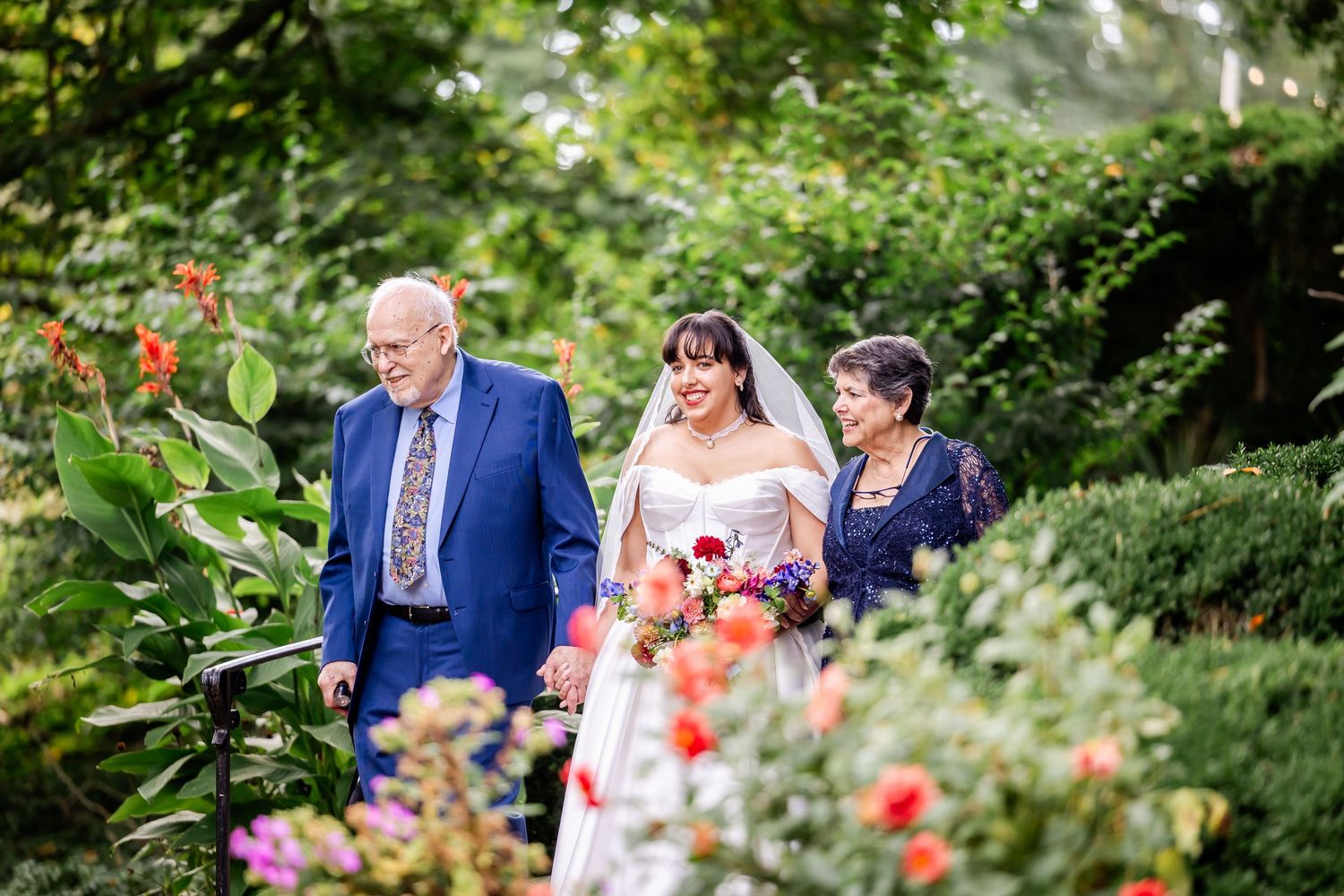 Bride walks through lush garden with elderly man in blue suit and woman in purple dress on her wedding day.