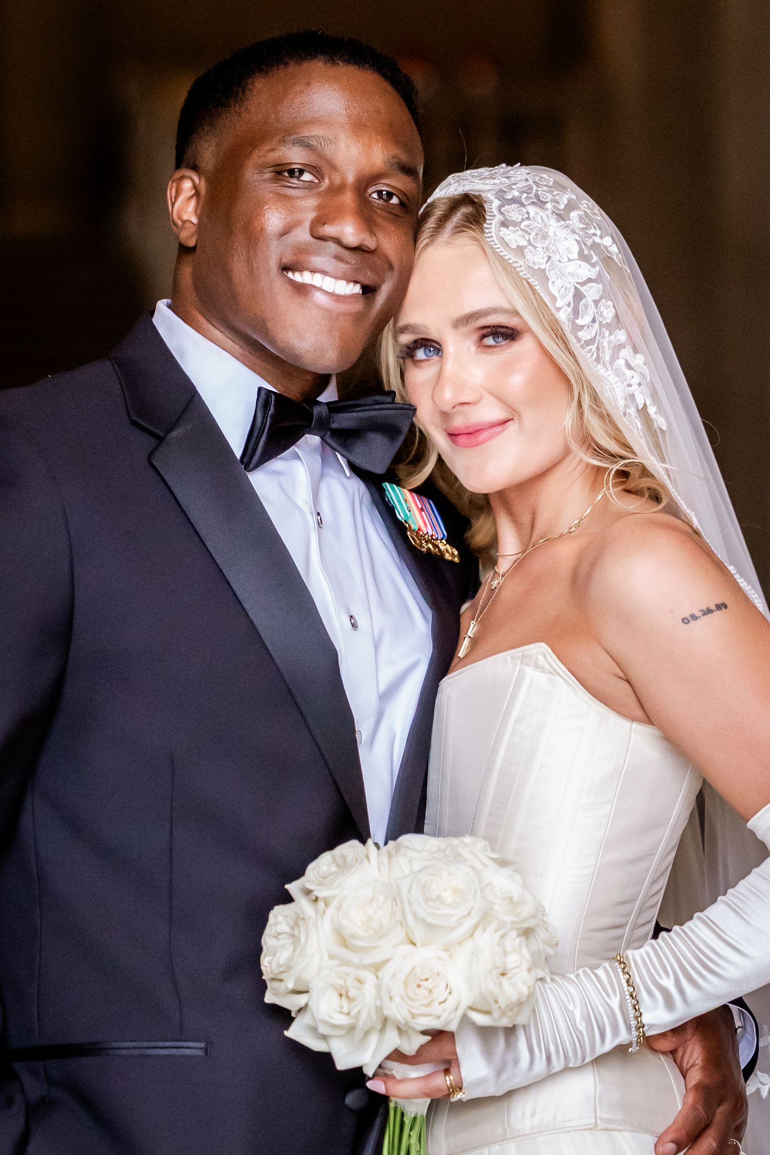 A smiling interracial couple poses in wedding attire, groom in tuxedo and bride in white gown holding roses.