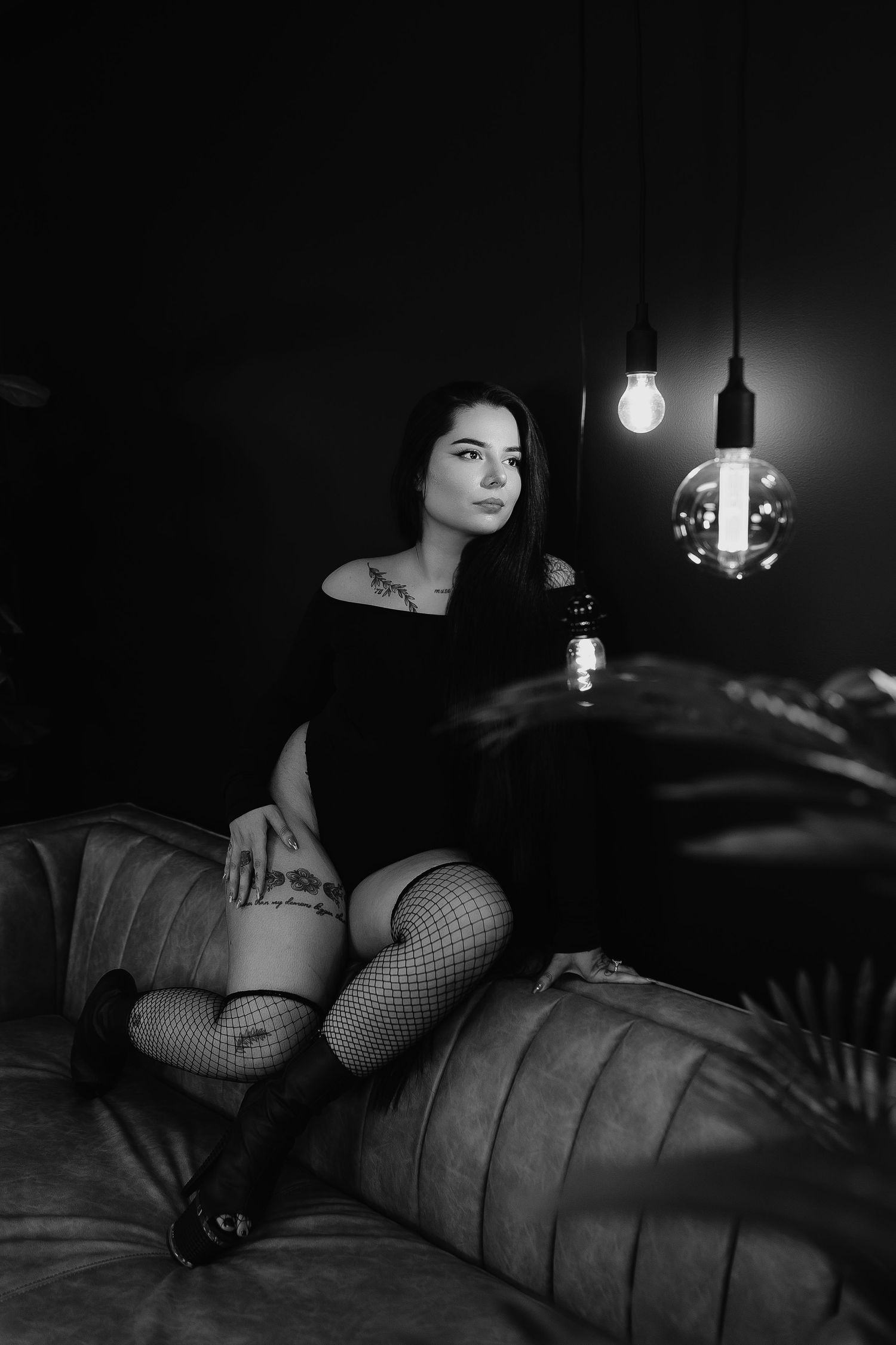 Black and white boudoir photo of a young woman in fishnet stockings sitting on a leather sofa near vintage Edison bulbs.