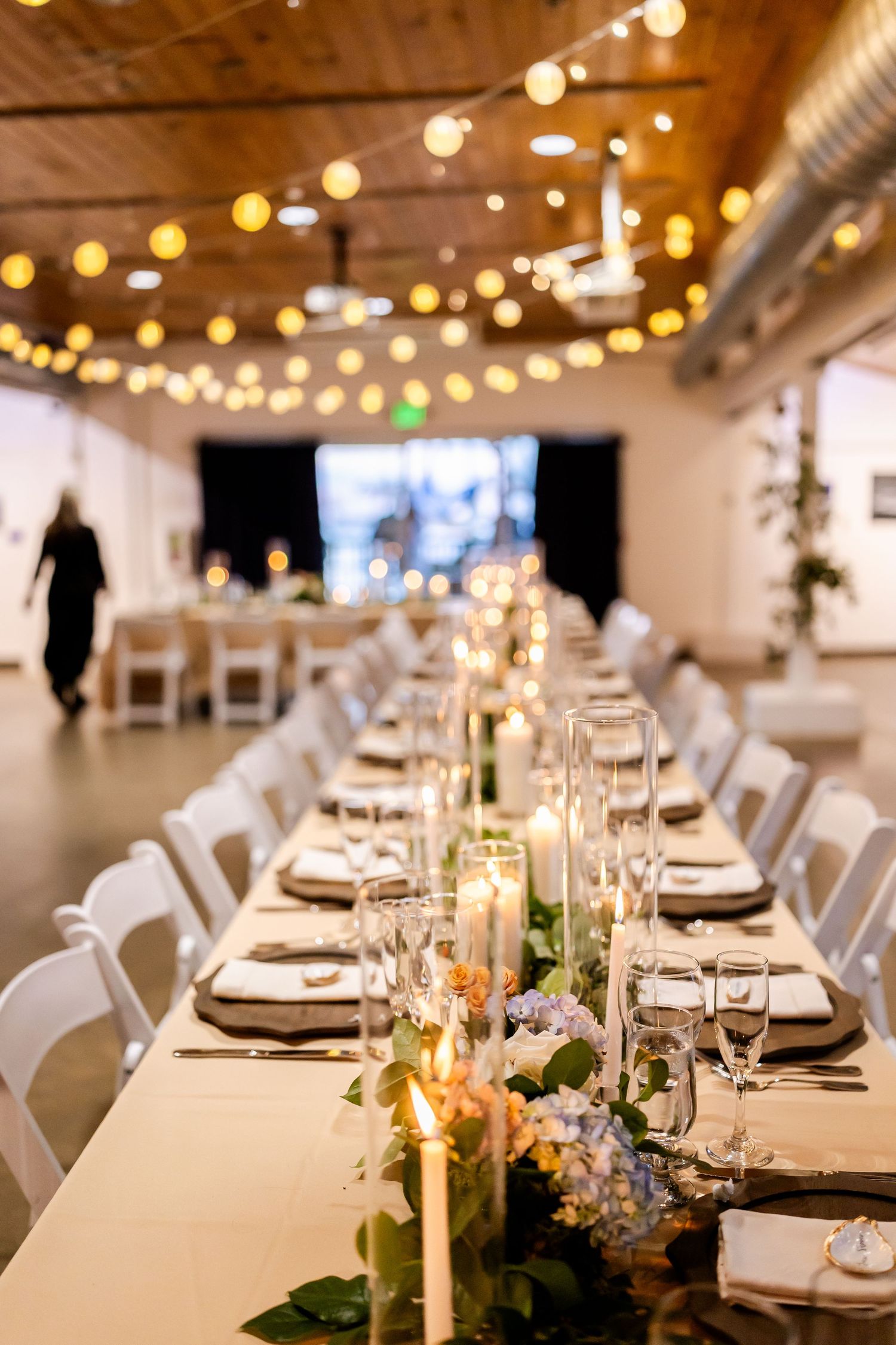 Elegant wedding reception hall with long tables, floral centerpieces, candles, and string lights overhead.