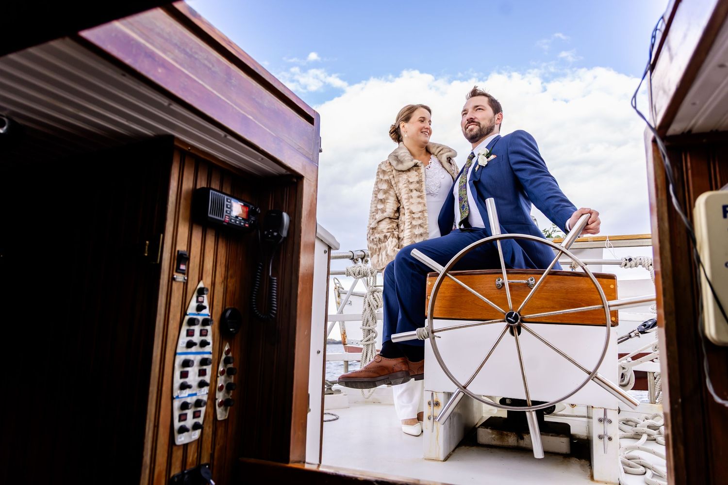 Couple in wedding attire standing at the helm of a wooden sailboat on a sunny day.