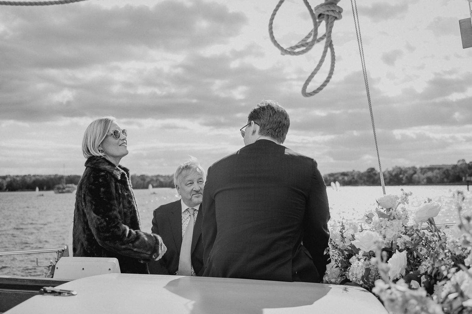 Three elegantly dressed guests conversing on a boat deck by a lake, captured in black and white photography.
