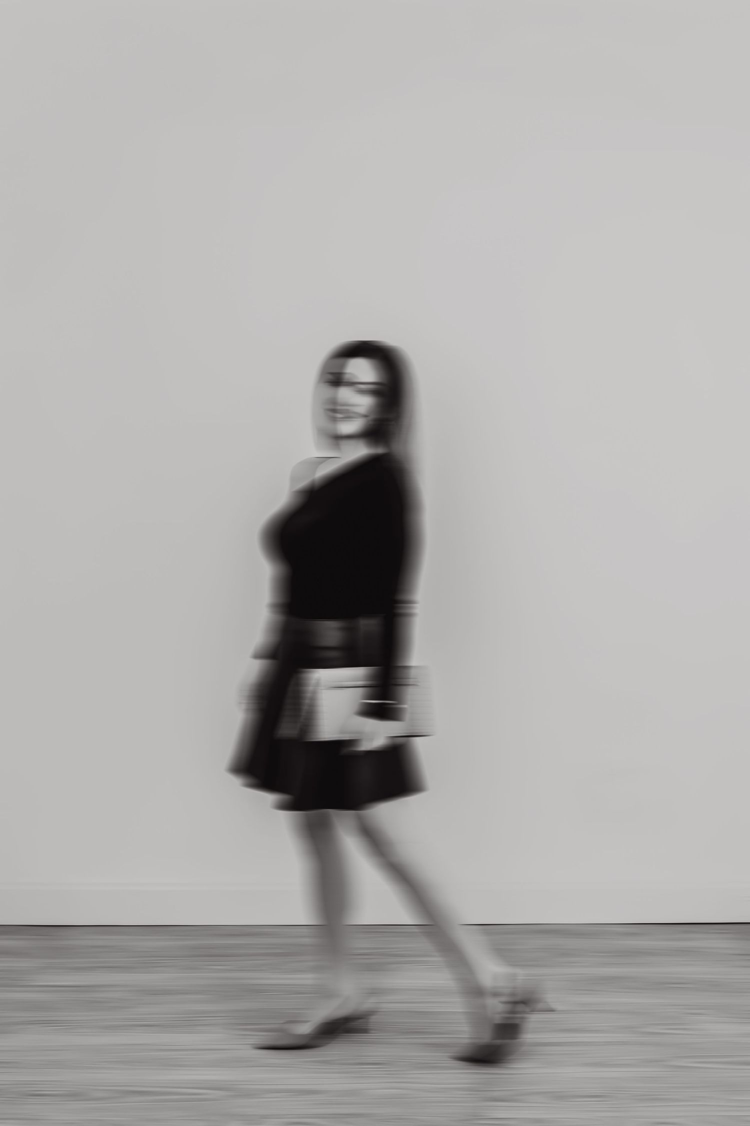 Black and white motion blur photo of a woman in a black dress walking confidently while holding a book.