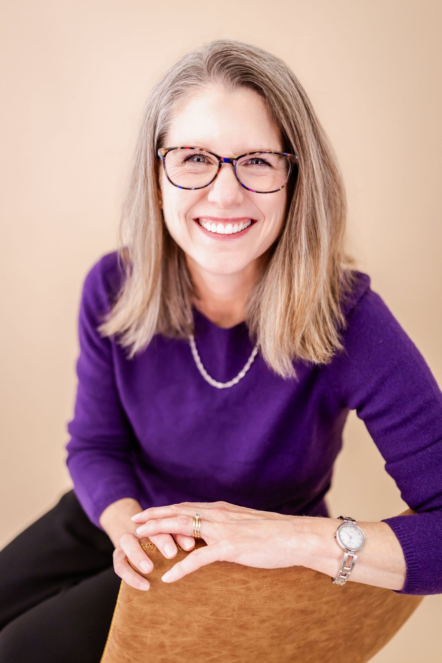Smiling woman with glasses and gray hair wearing a purple sweater and pearl necklace, seated against a beige background.