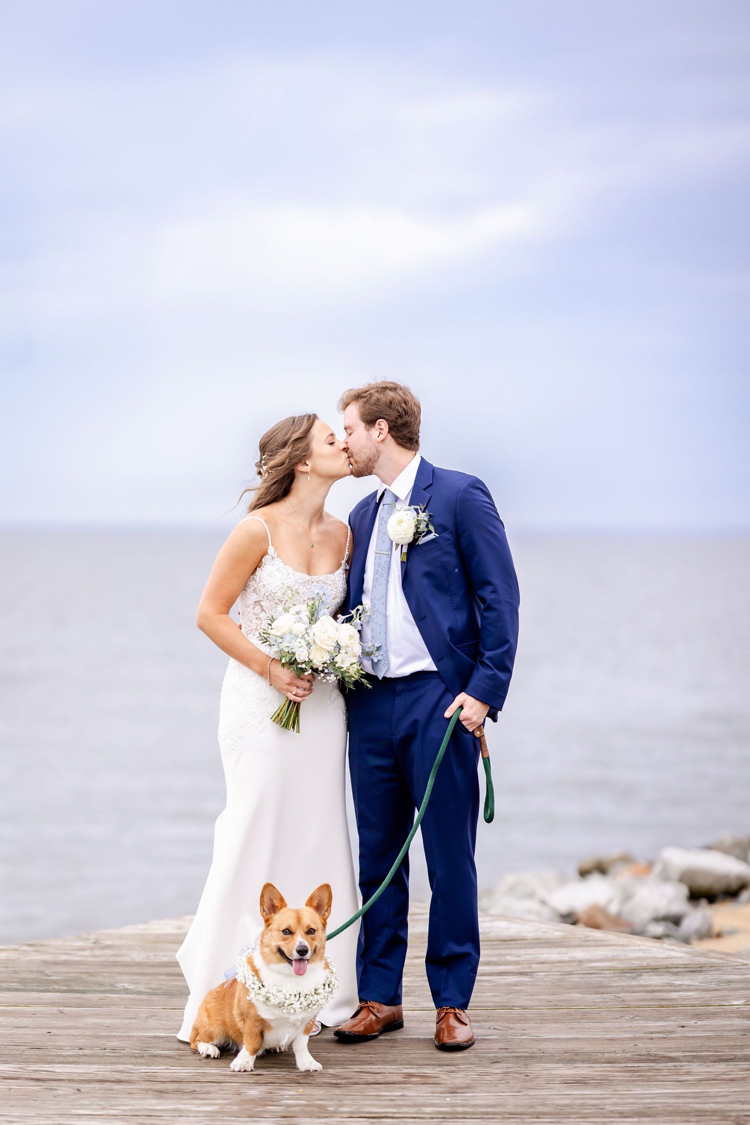 Wedding couple posing with their corgi dog wearing a floral collar on a wooden dock.