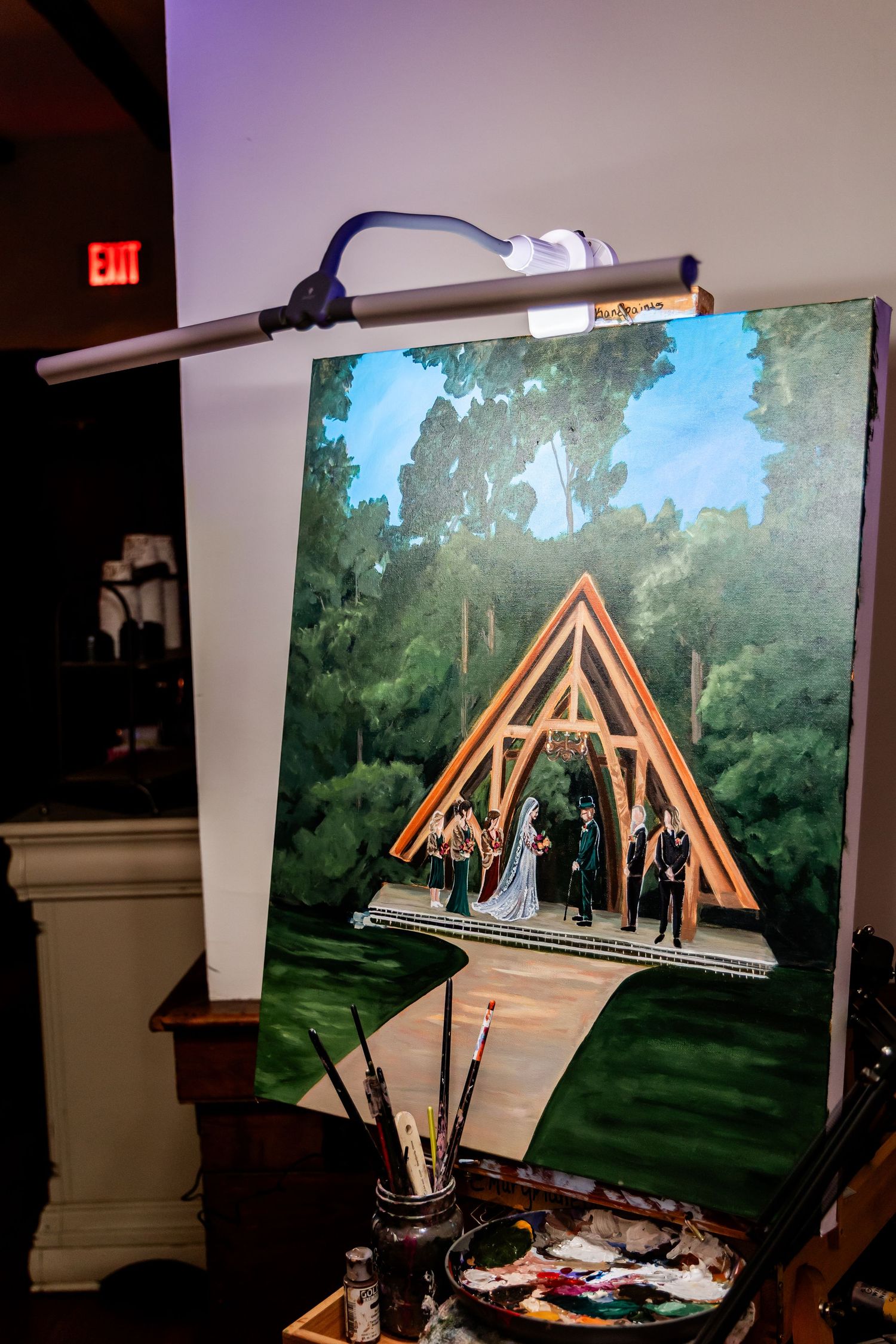 A painting on an easel shows a wooden A-frame wedding arch in a lush outdoor setting.