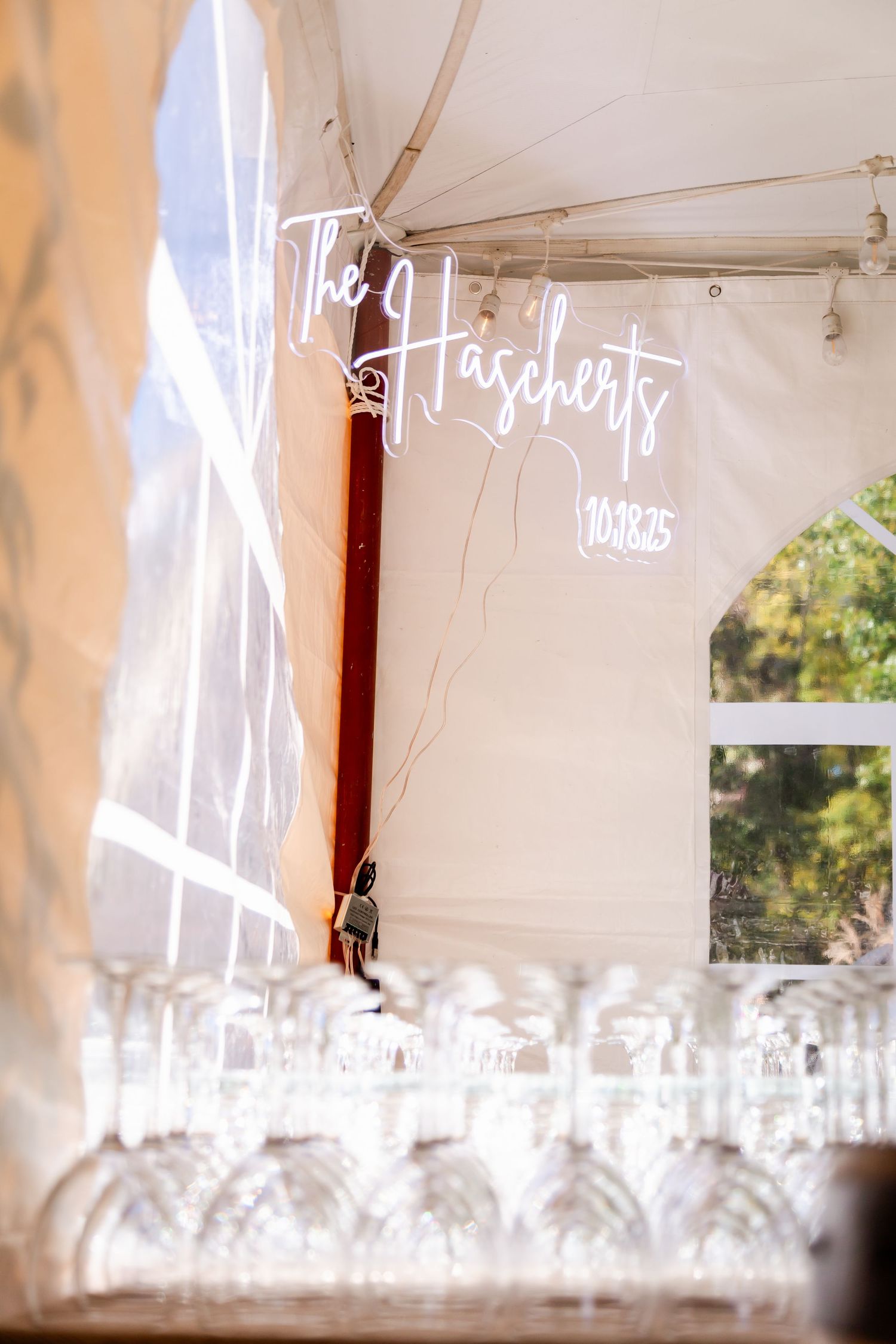 A row of empty wine glasses lined up under a white tent canopy with cursive text visible above.