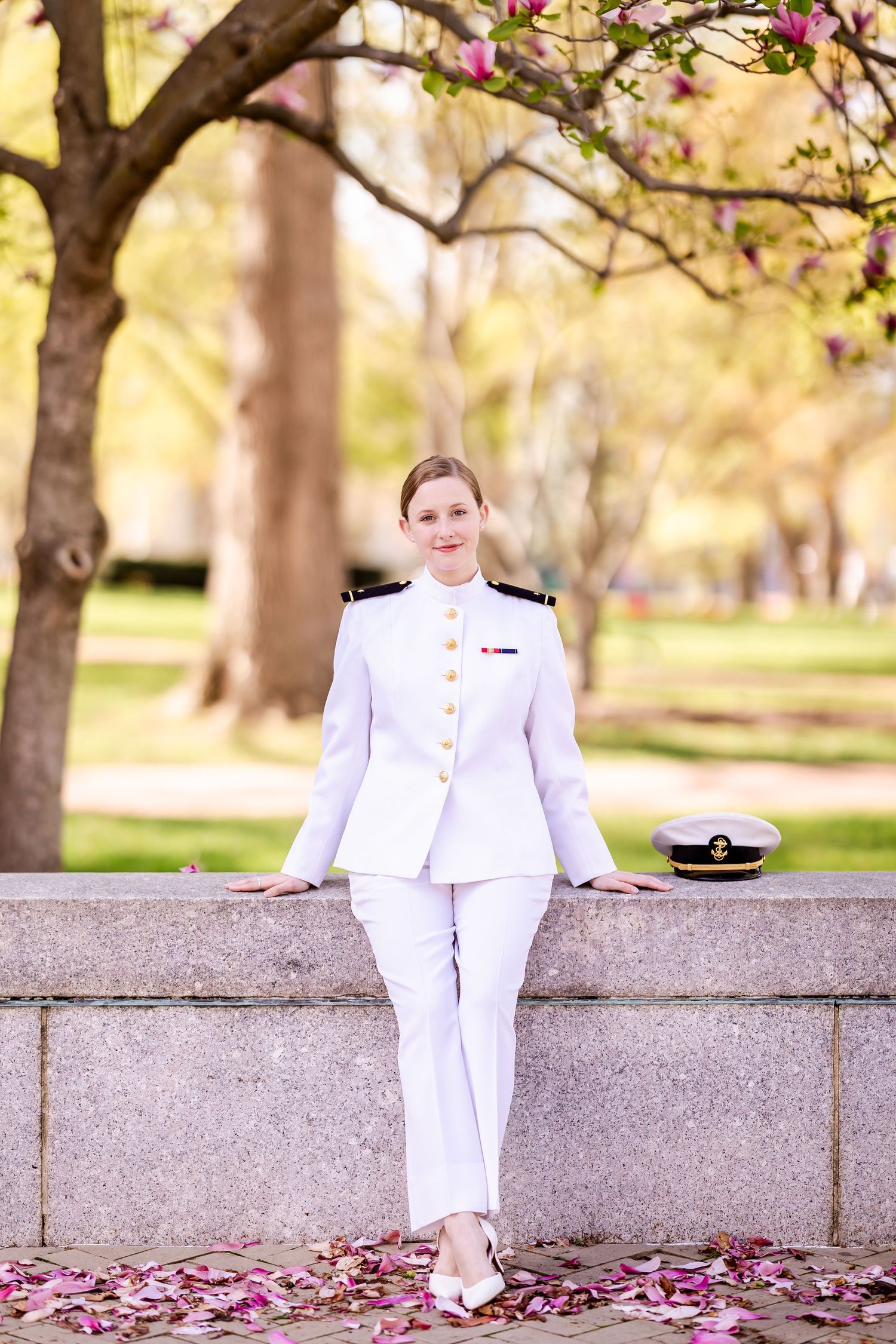 A person in a formal white military dress uniform stands against a stone wall under blossoming cherry trees in springtime.