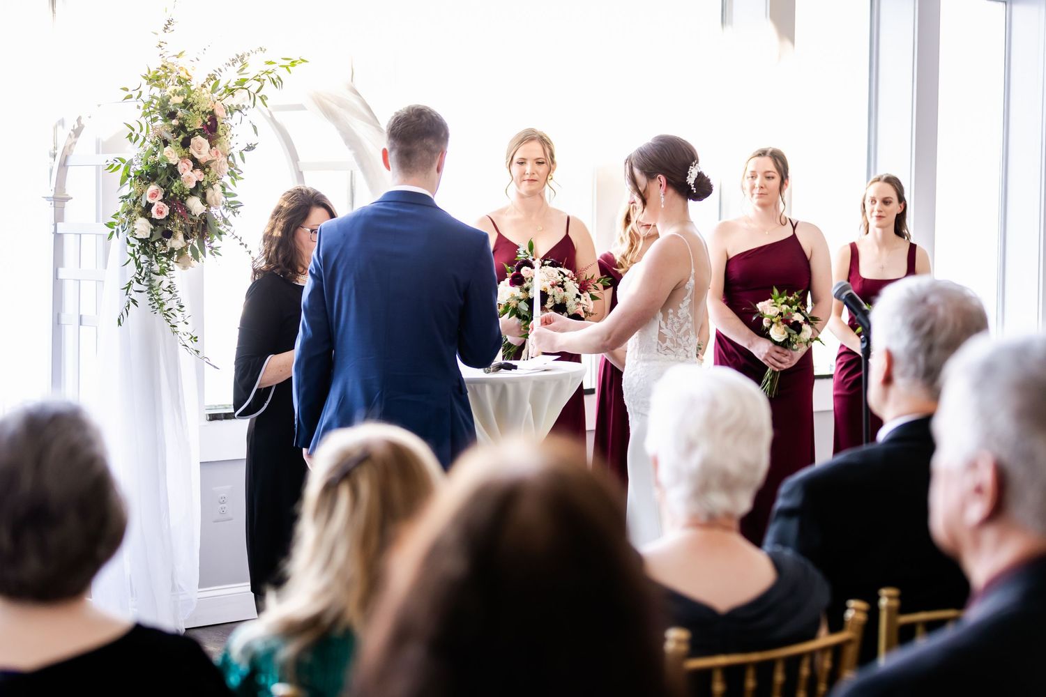 Indoor wedding ceremony with burgundy-clad attendants and floral arch decorations.