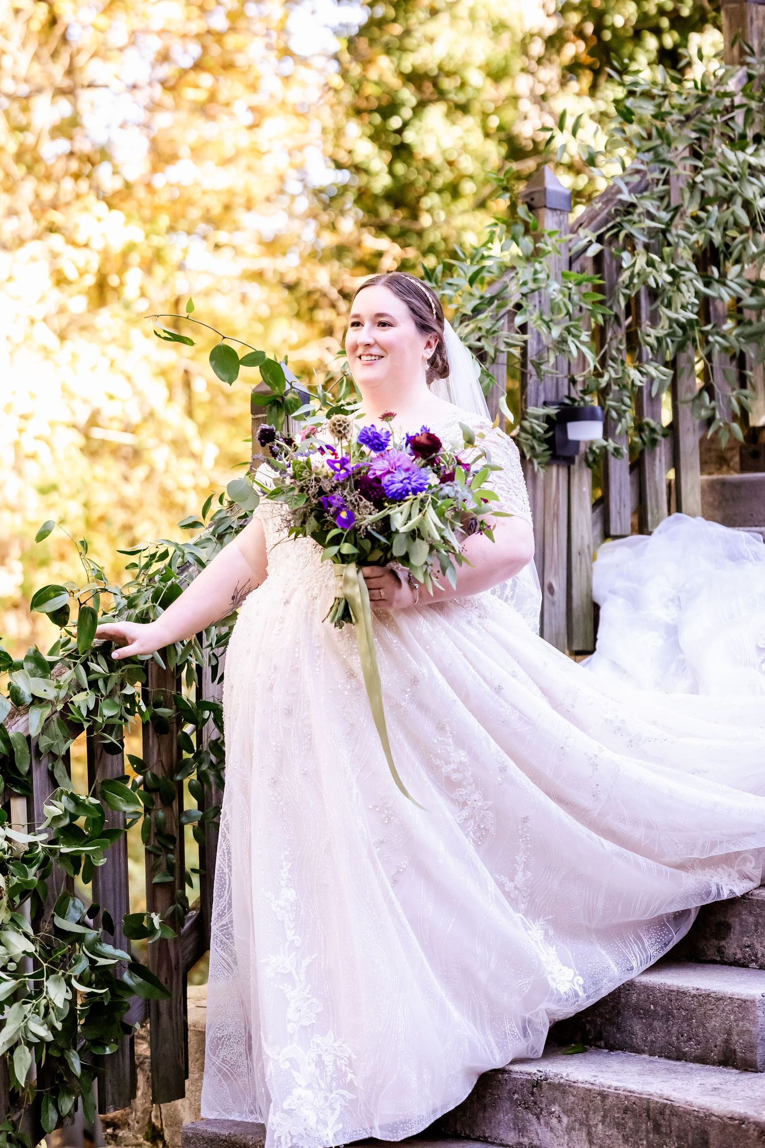 Bride in lace gown holds purple bouquet while posing on garden steps with flowing train.