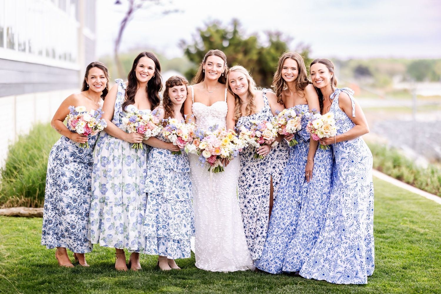 Bridesmaids in blue floral dresses pose together on grassy lawn.