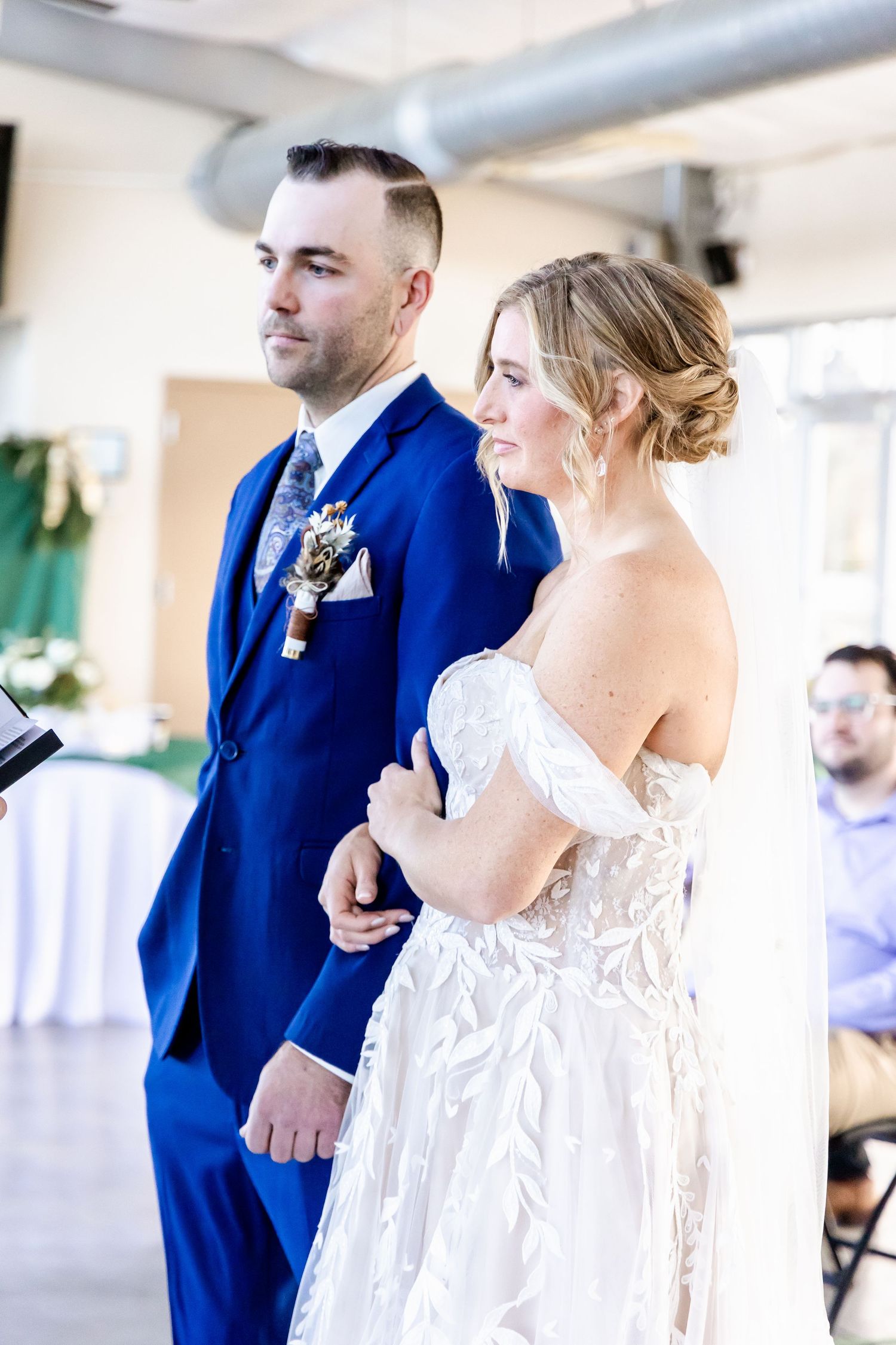 A couple stands at the altar during their wedding ceremony, with the bride in white lace and groom in royal blue suit.