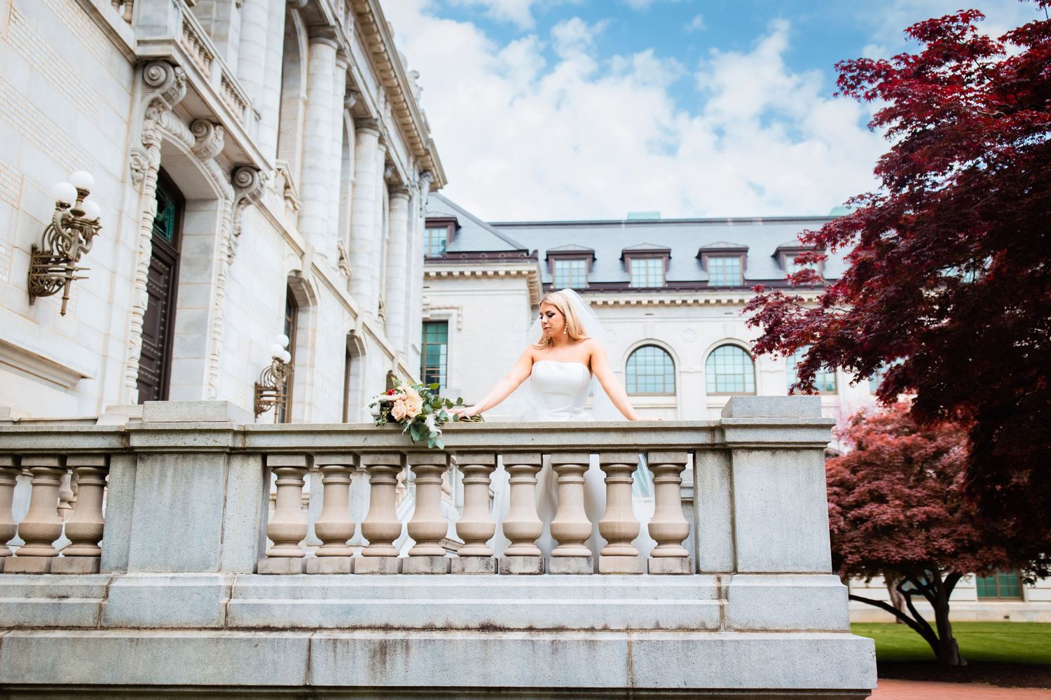 A white wedding dress flows over an elegant stone balustrade at a classic architectural venue with red maple trees.