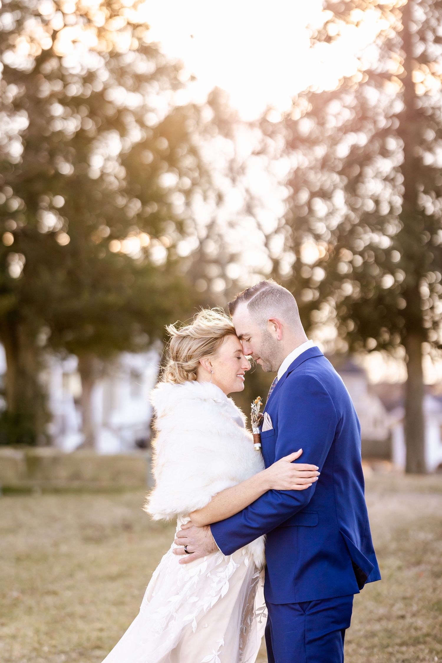 Romantic wedding couple embraces outdoors at sunset in blue suit and white dress with fur wrap.