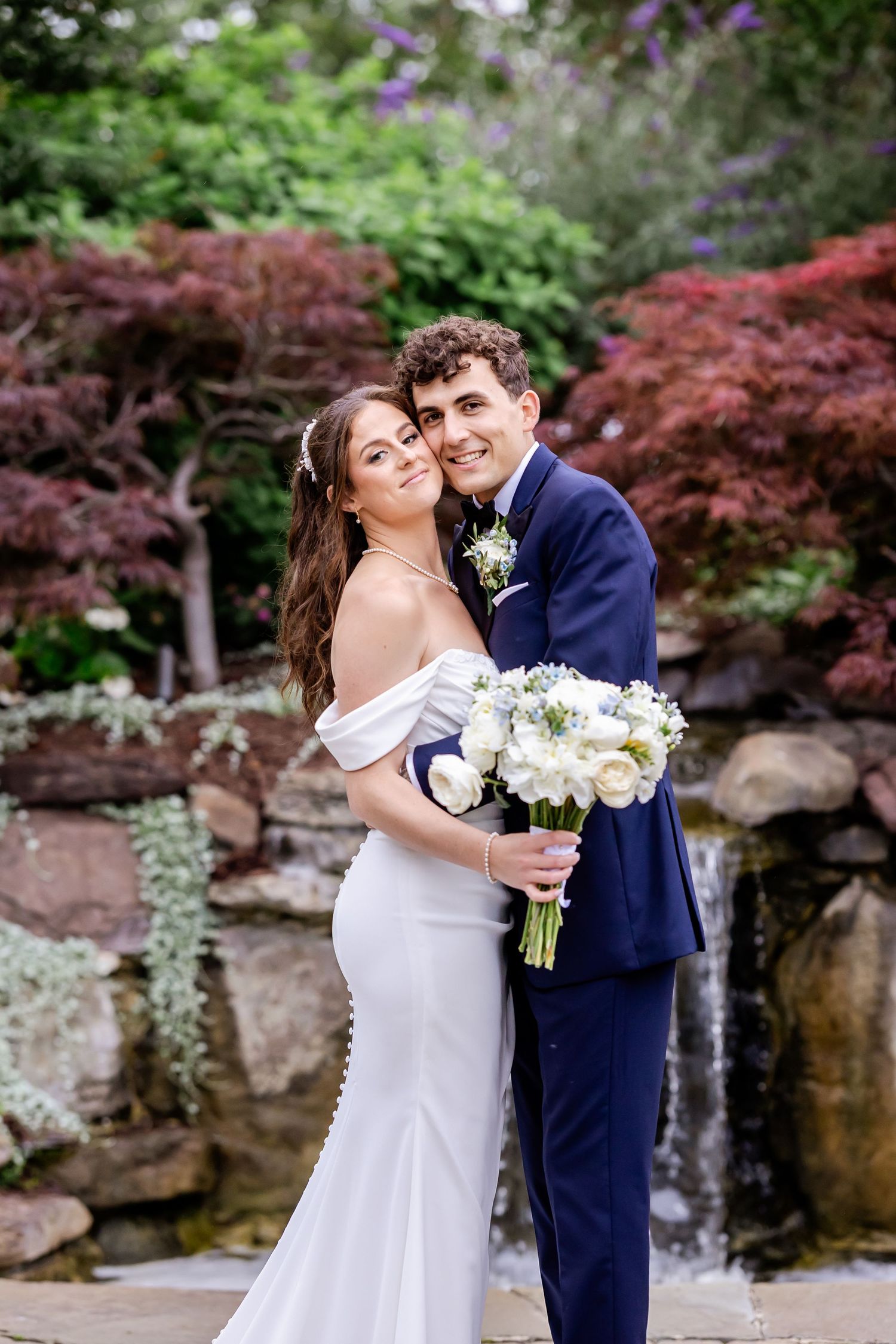 Romantic wedding couple poses in garden setting with red maple trees and white floral bouquet.