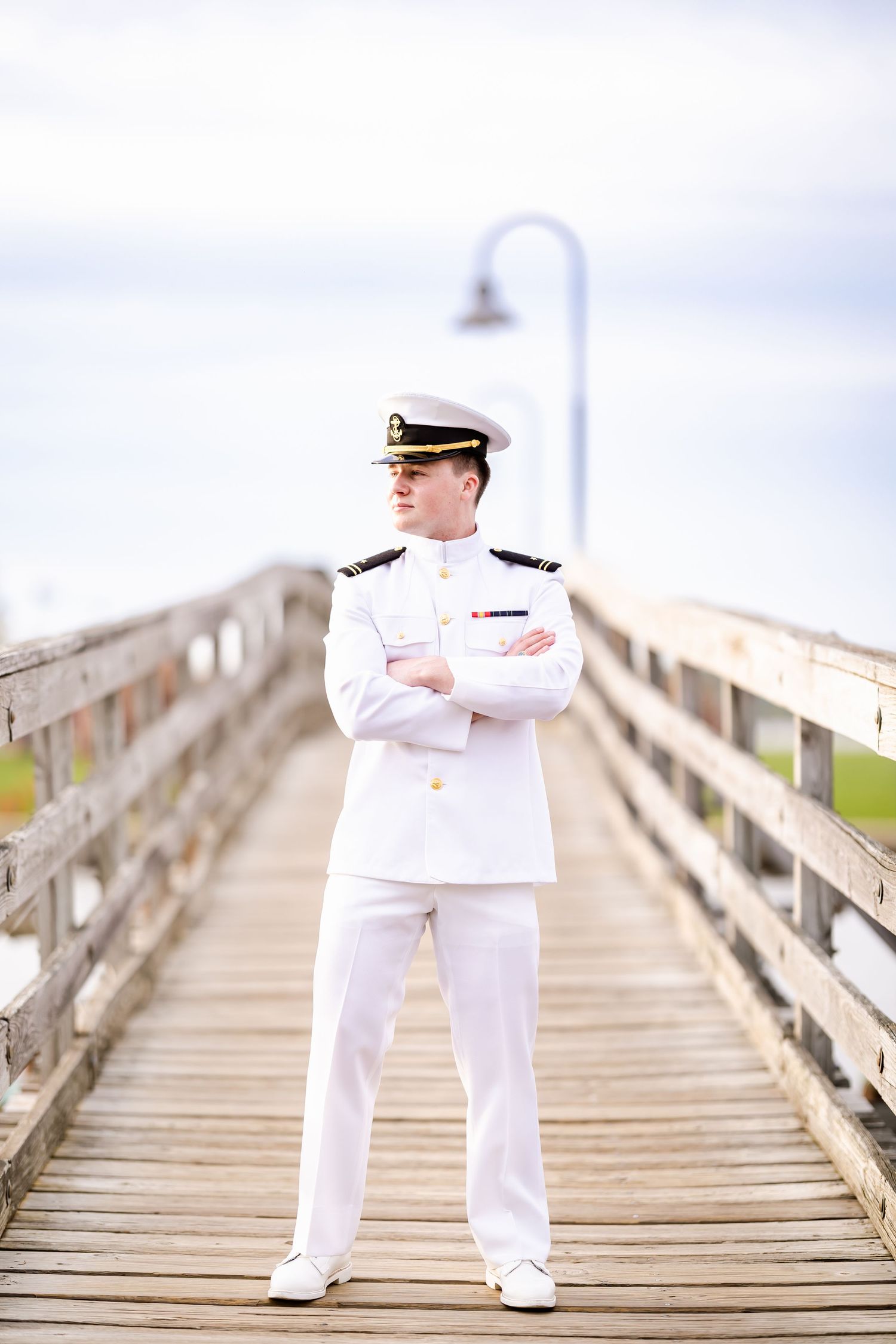Naval officer in white dress uniform stands confidently on wooden pier with arms crossed.