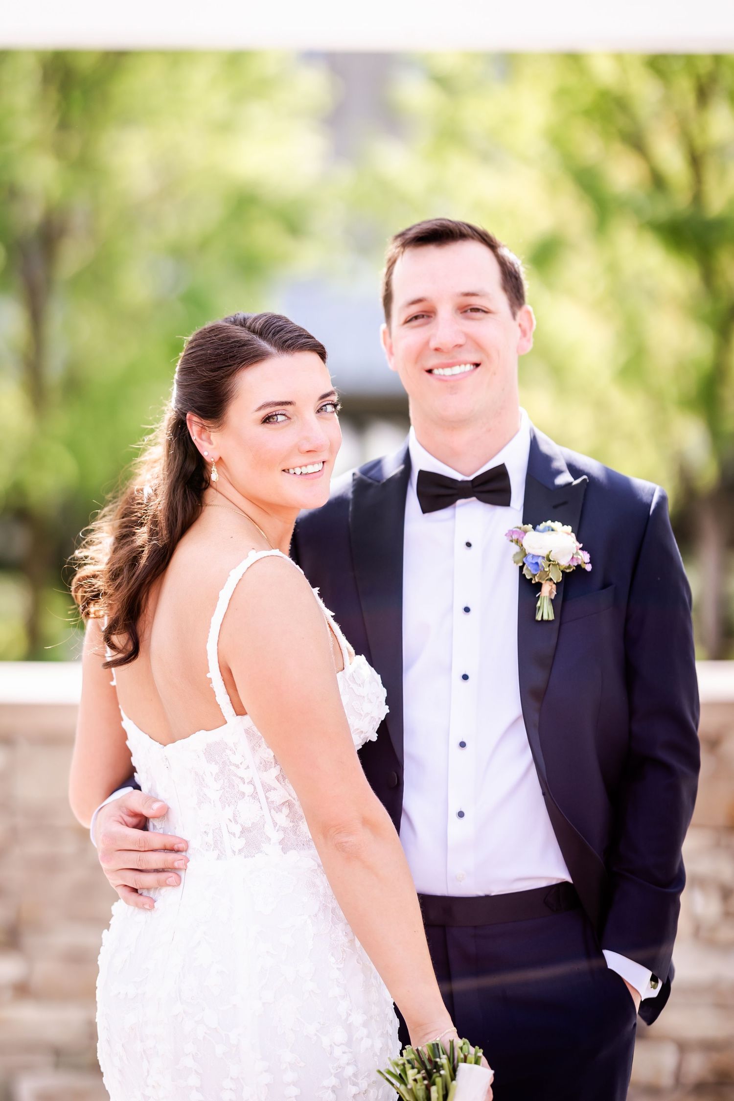 A bride in a white dress and groom in a black tuxedo pose together outdoors for their wedding portrait.