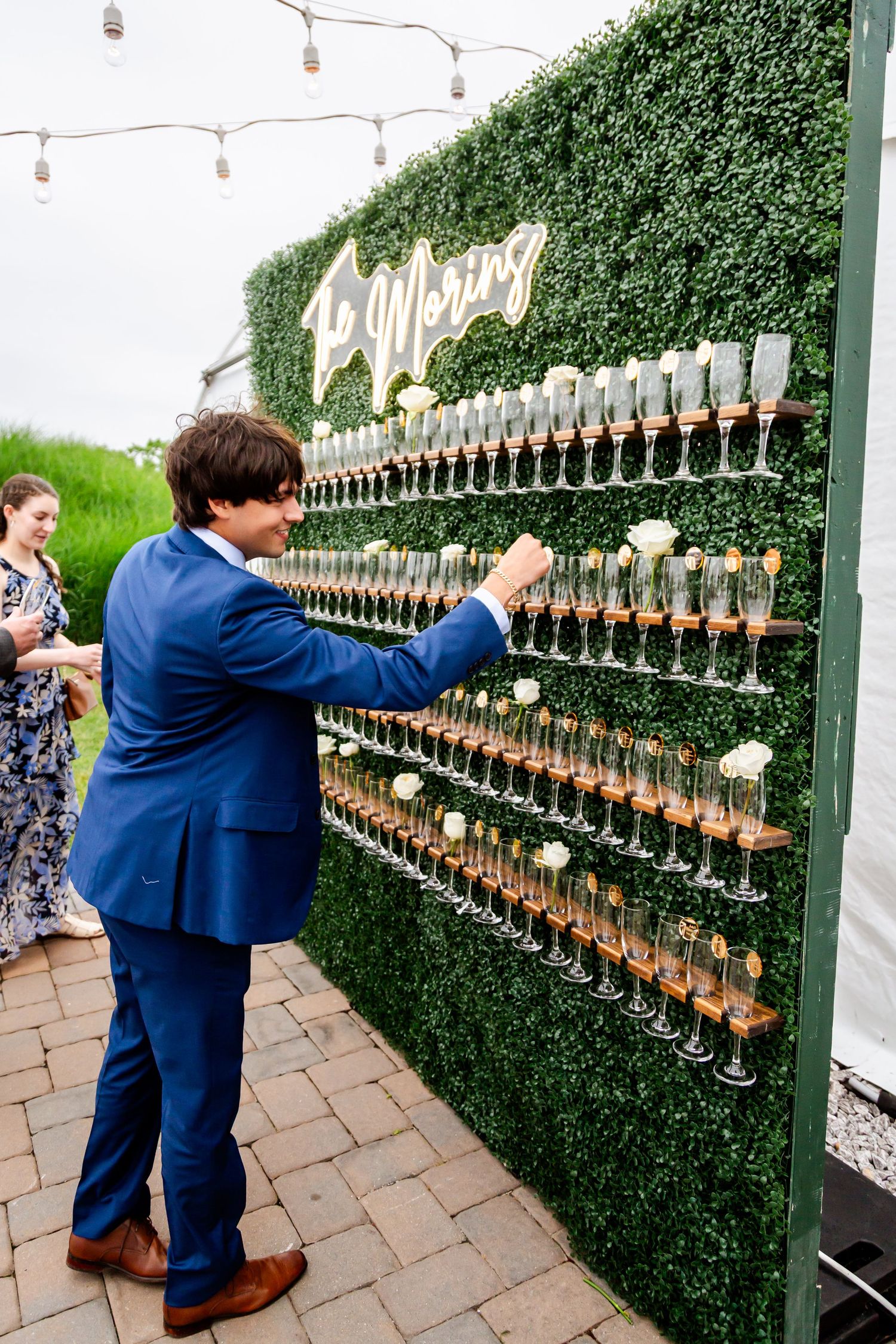 A wall display of champagne glasses decorated with greenery and string lights at a wedding reception.