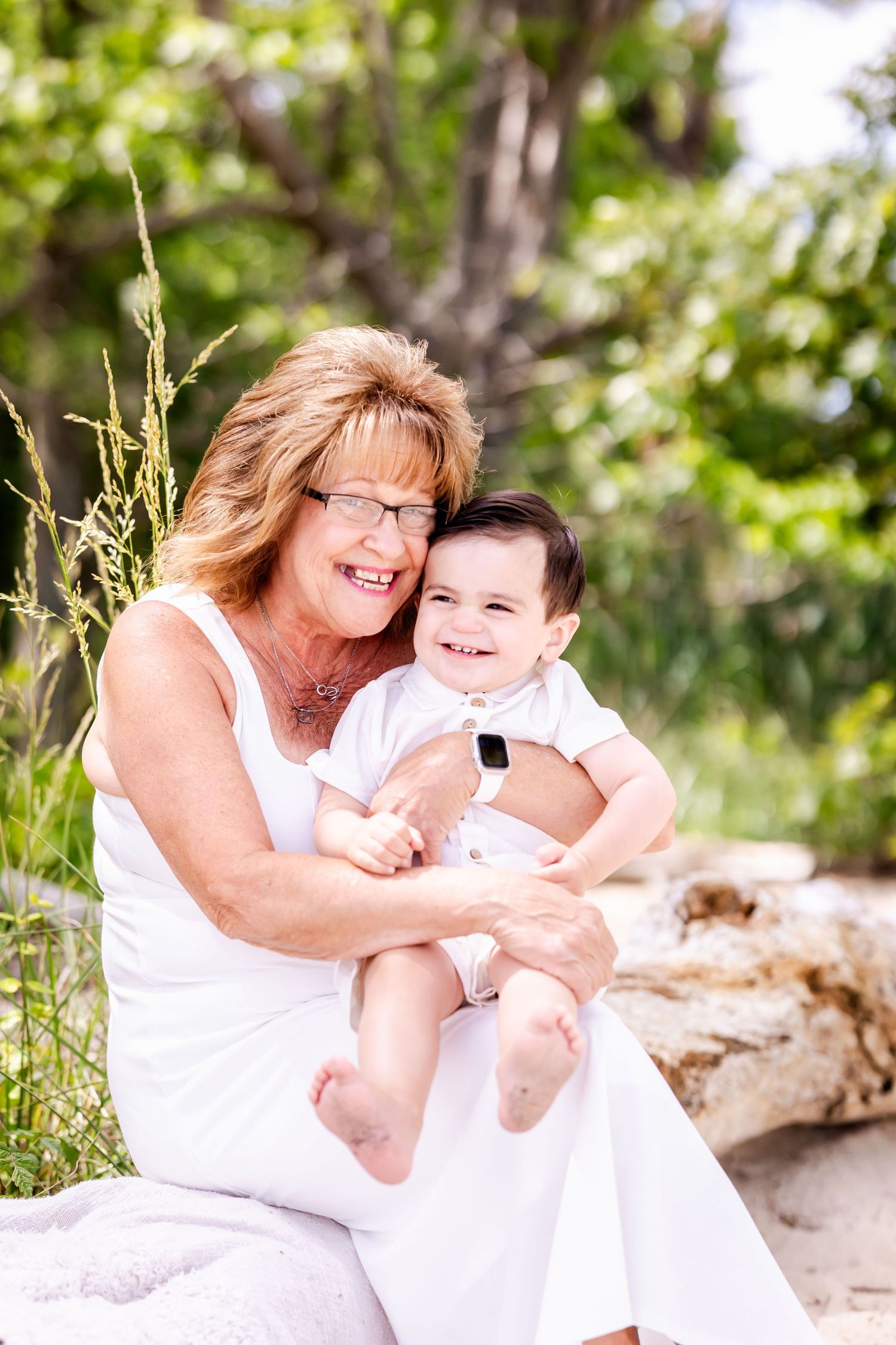 A woman in a white dress sits outdoors holding a happy baby against a nature background.