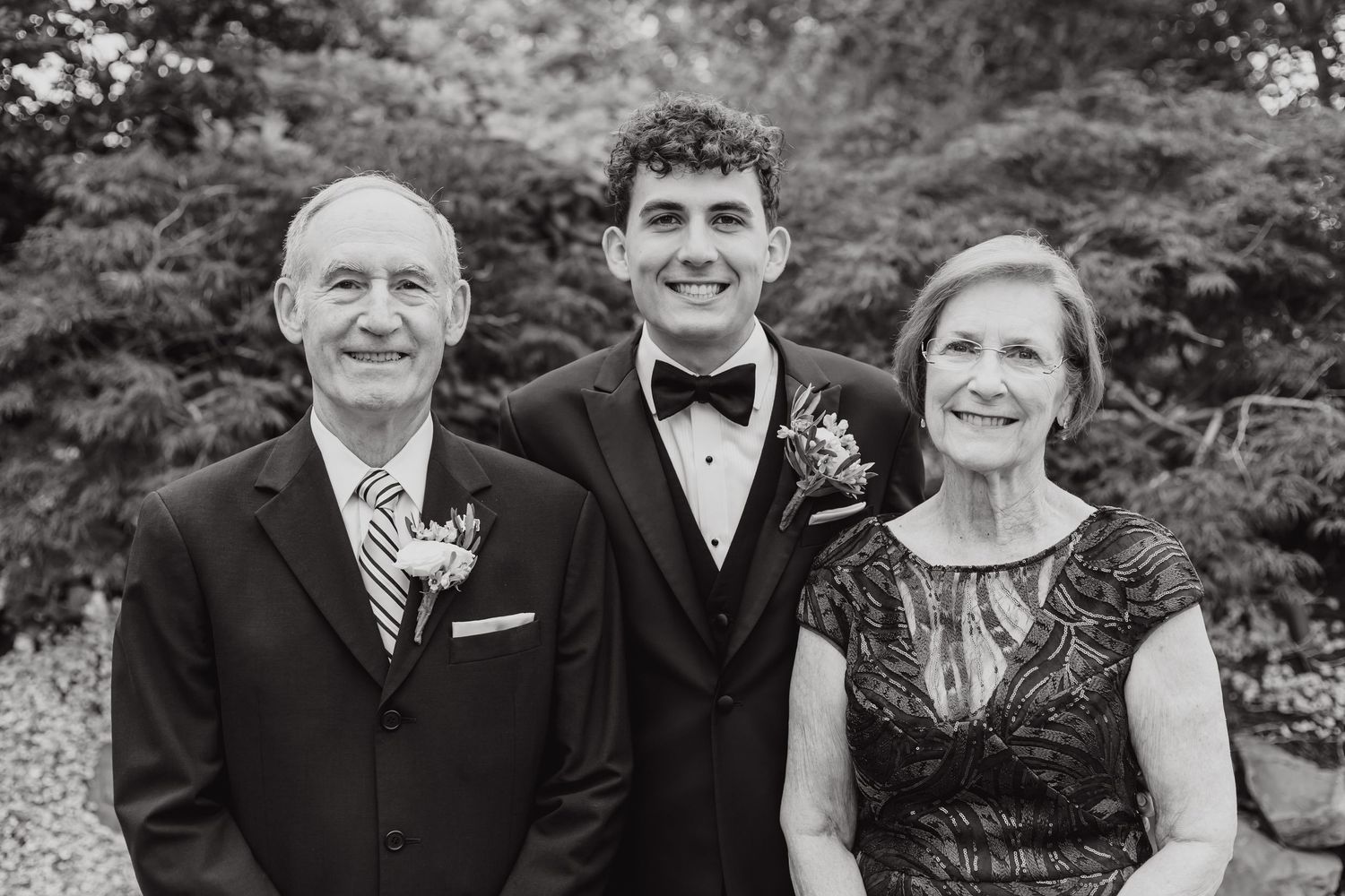 Formal black and white portrait of wedding party members outdoors with greenery backdrop.