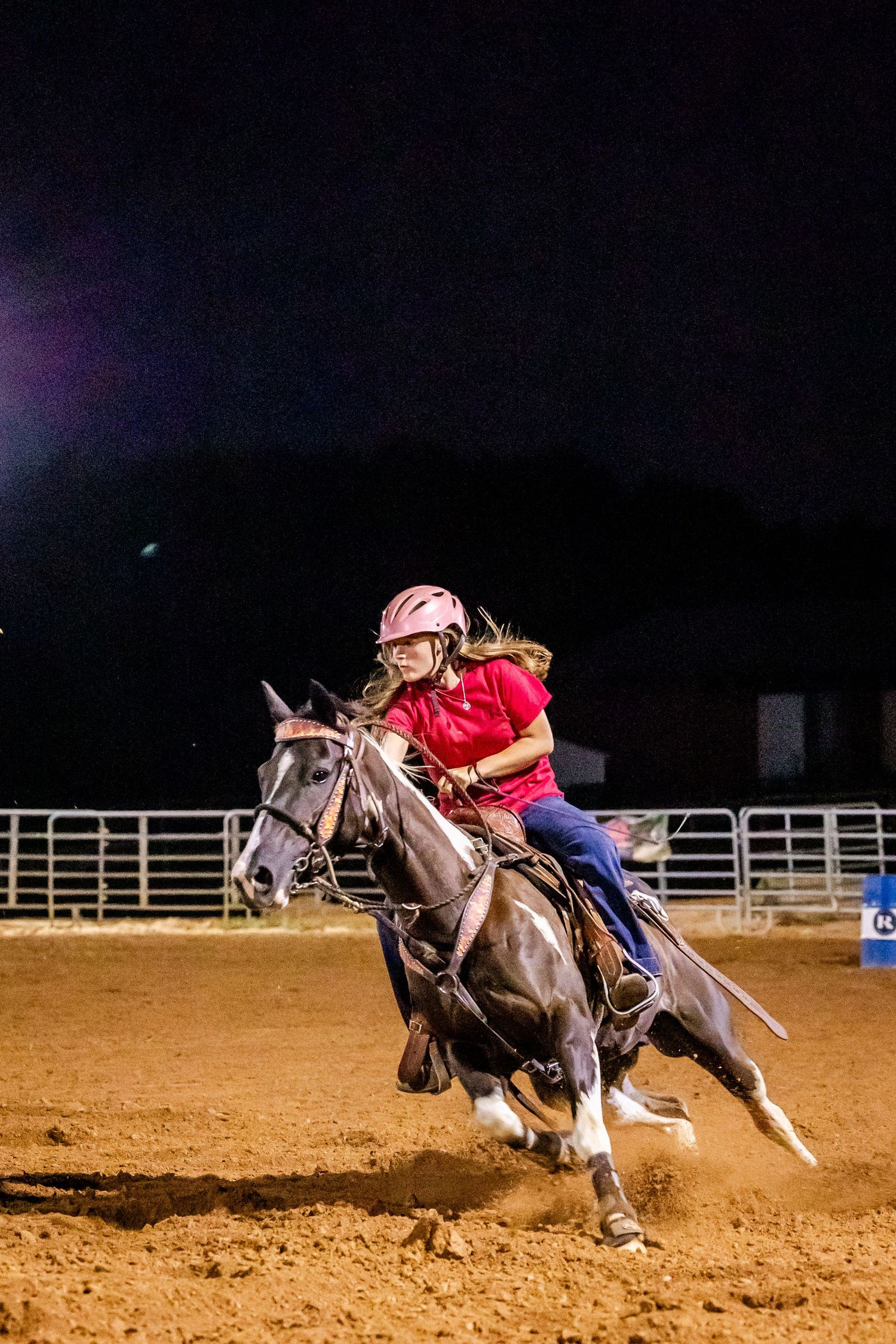 Nighttime barrel racing competition with rider guiding horse through fast-paced turns in rodeo arena.