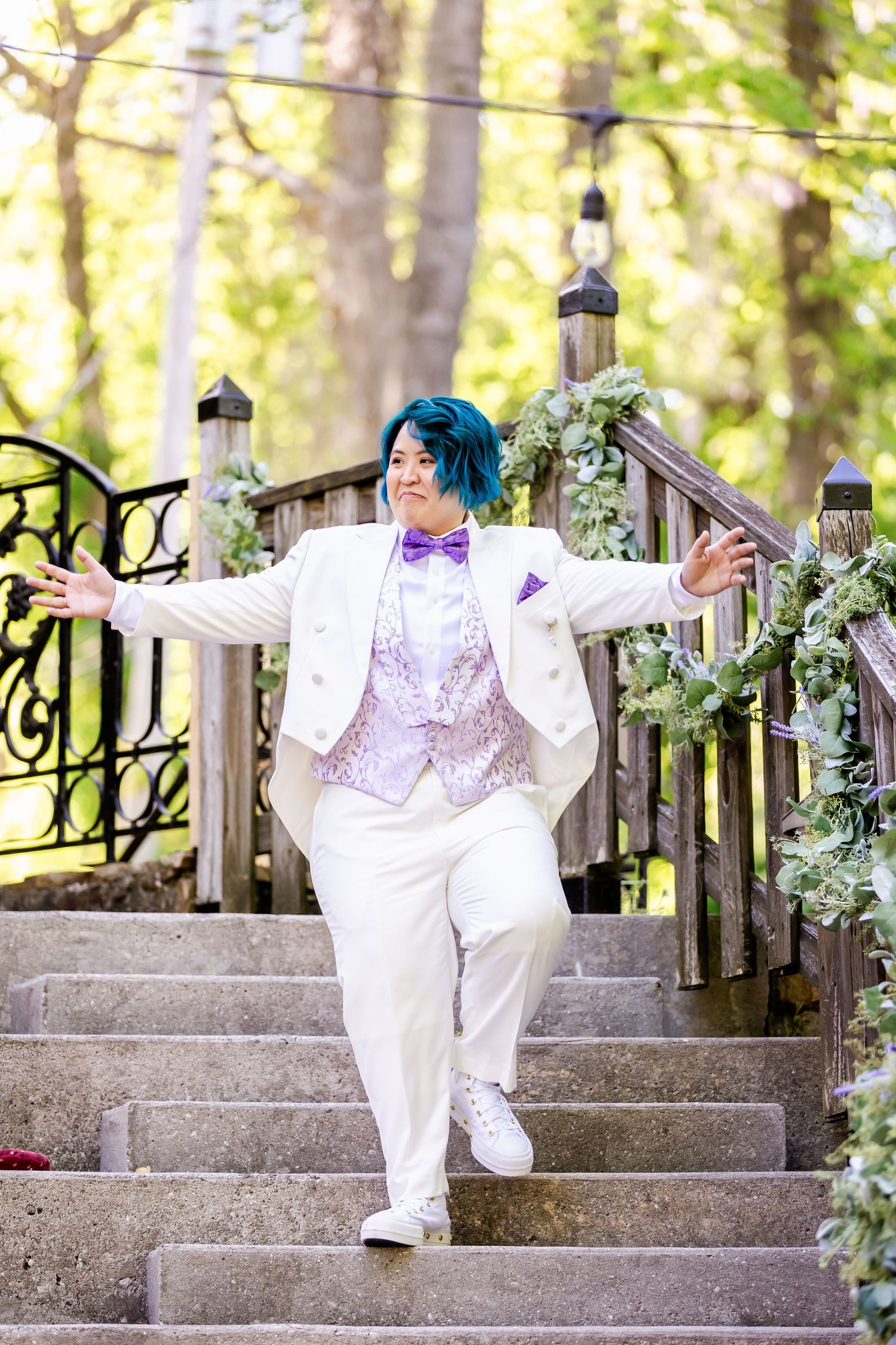 Person in white suit and purple bowtie poses dramatically on outdoor decorated staircase.