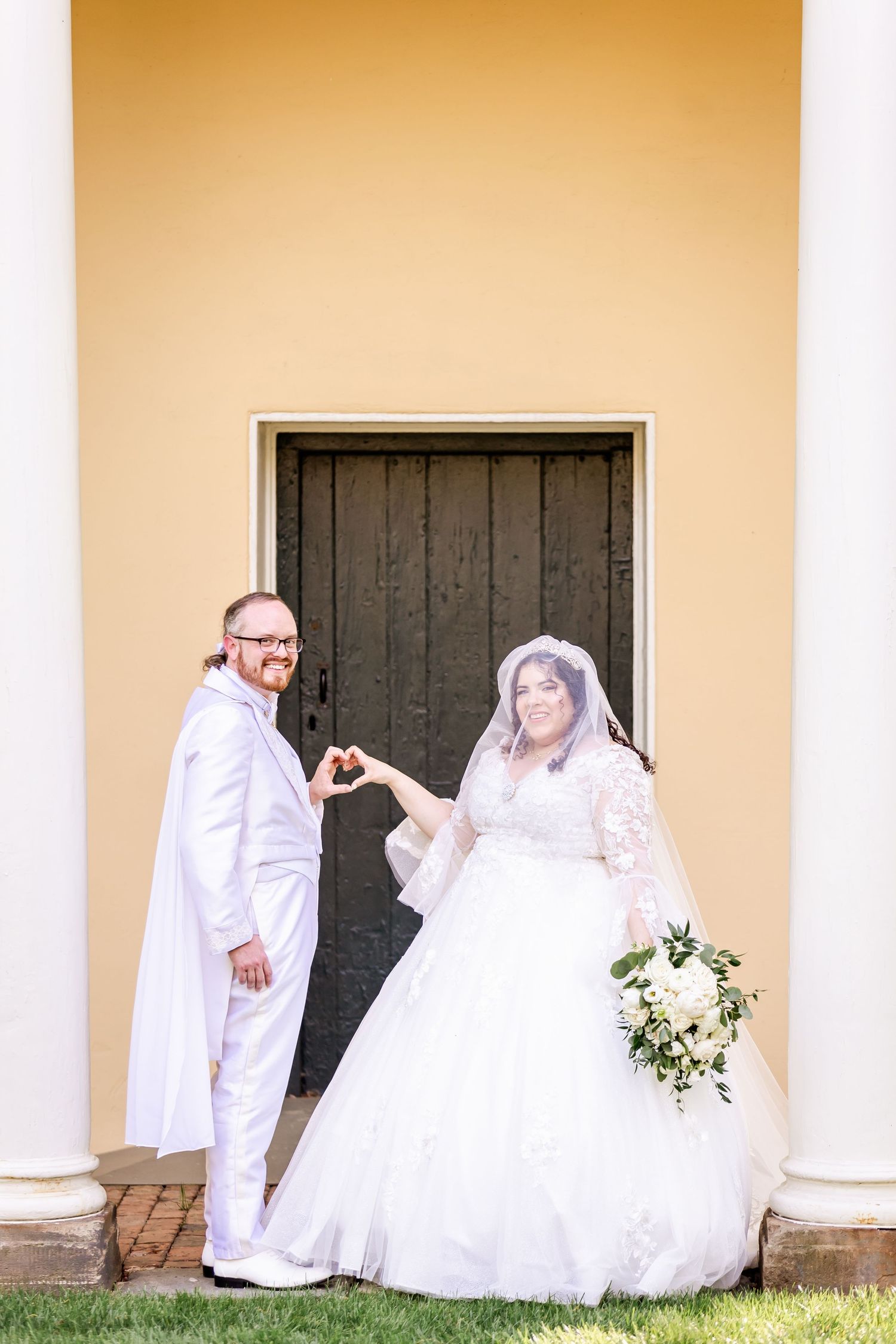 Wedding couple poses playfully by wooden door of yellow colonial building with white columns.