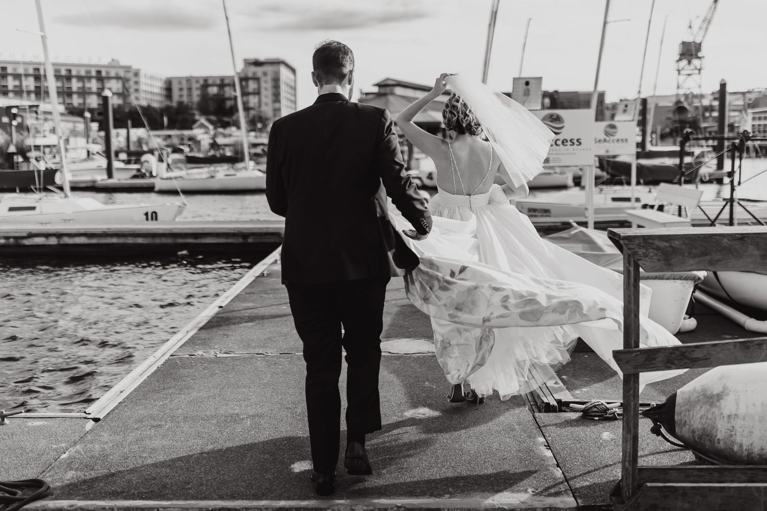 A black and white photo of newlyweds walking along a marina dock with flowing veil.