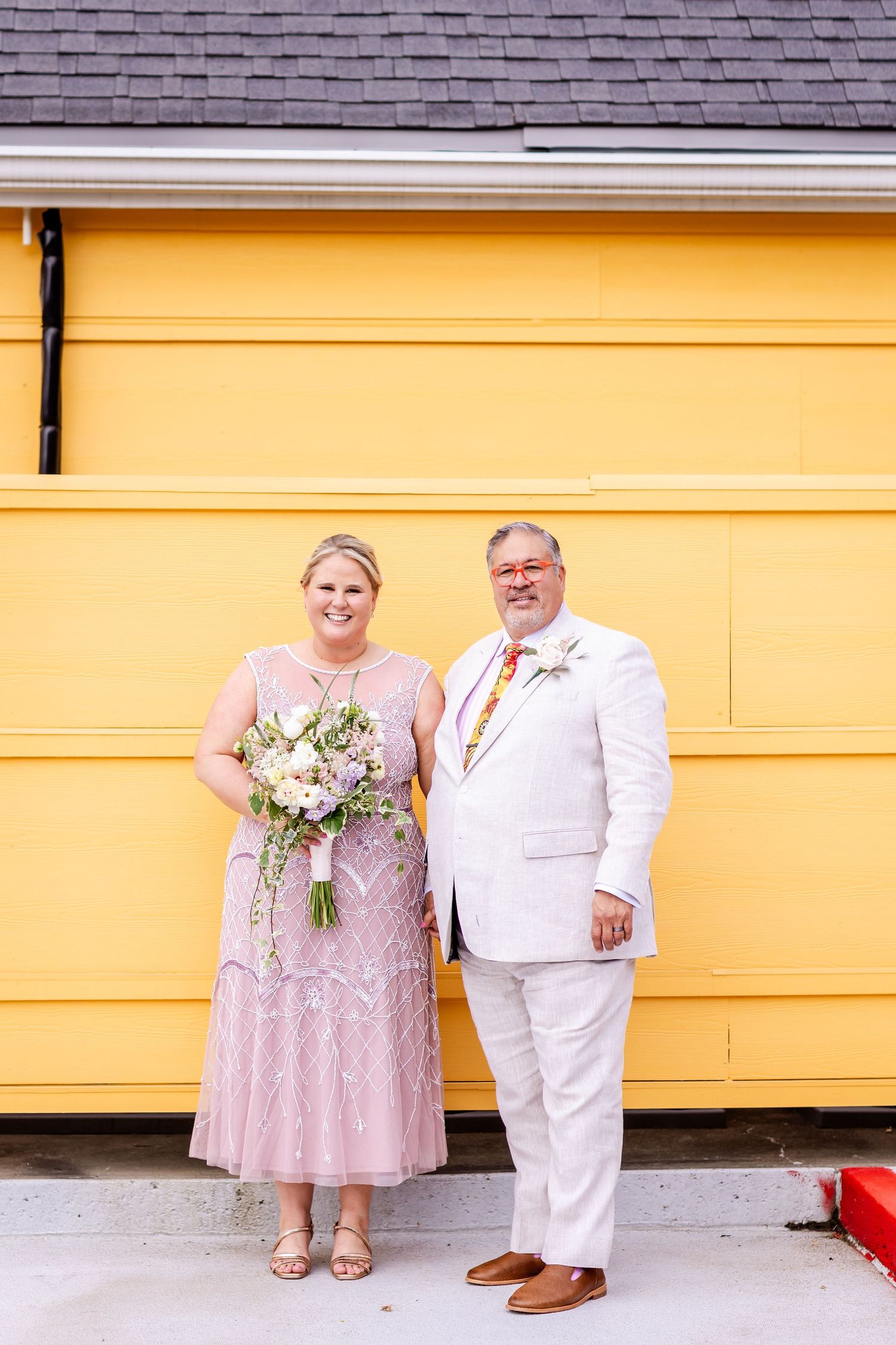 A formal couple poses against a vibrant yellow building wearing matching pink and white outfits.