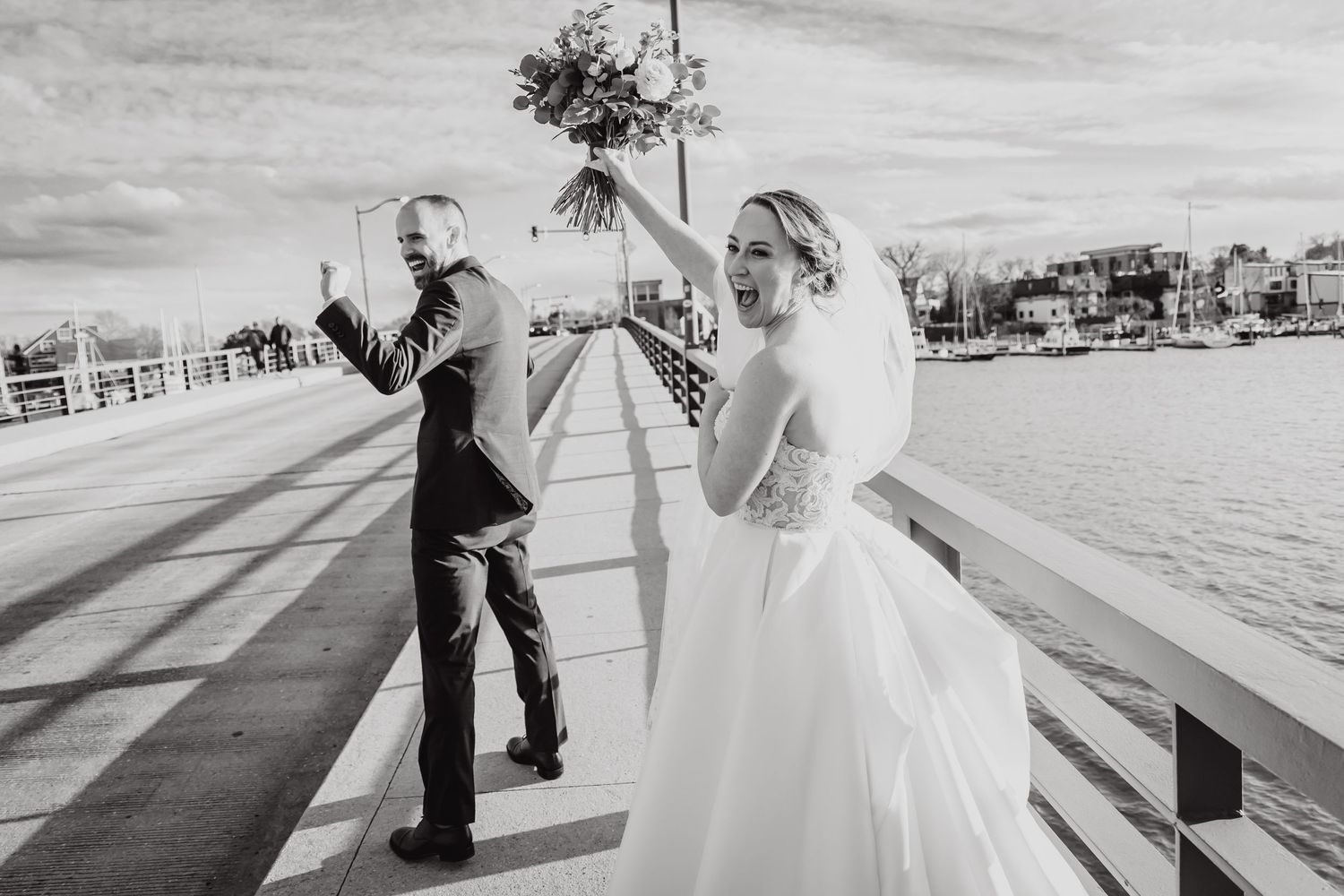 A wedding couple enjoys a playful candid moment on a waterfront boardwalk during their black and white photo session.