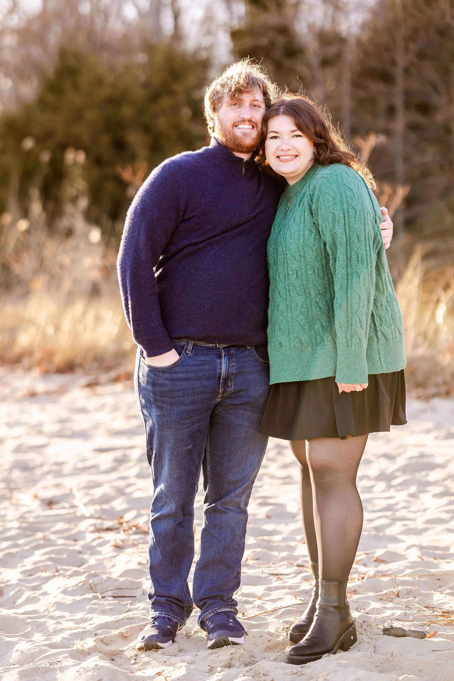 Couple wearing navy and green sweaters poses together on beach during sunset.