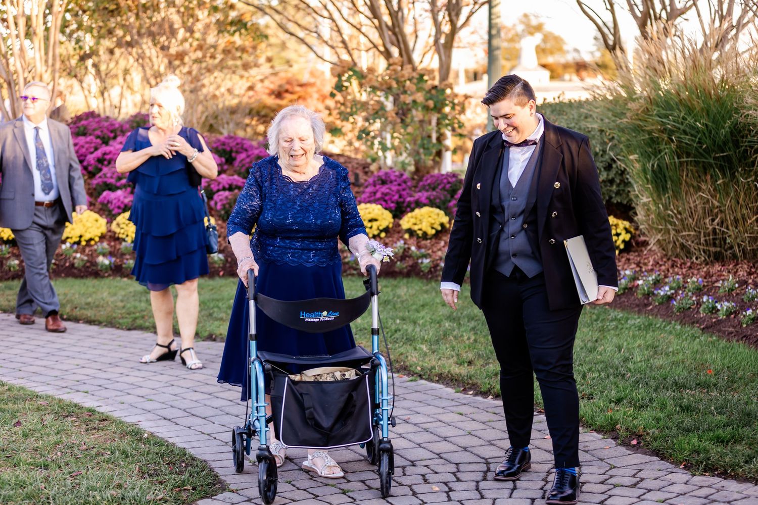 Formal event attendees stroll along a stone pathway surrounded by autumn gardens and flowers.