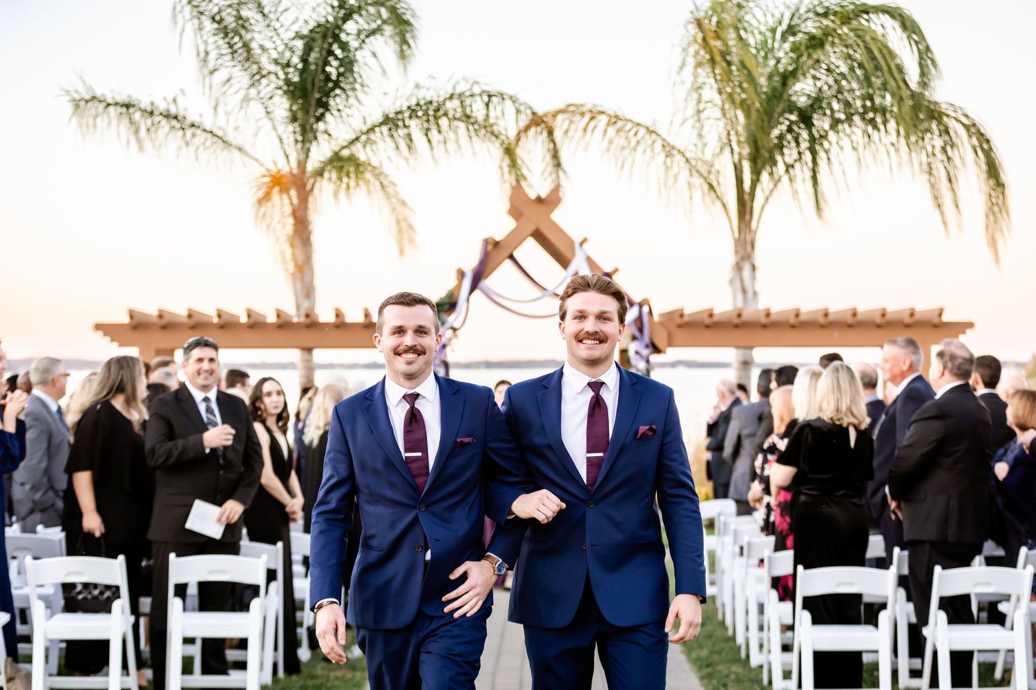 Wedding party members in navy suits walk down aisle under palm trees at outdoor ceremony.