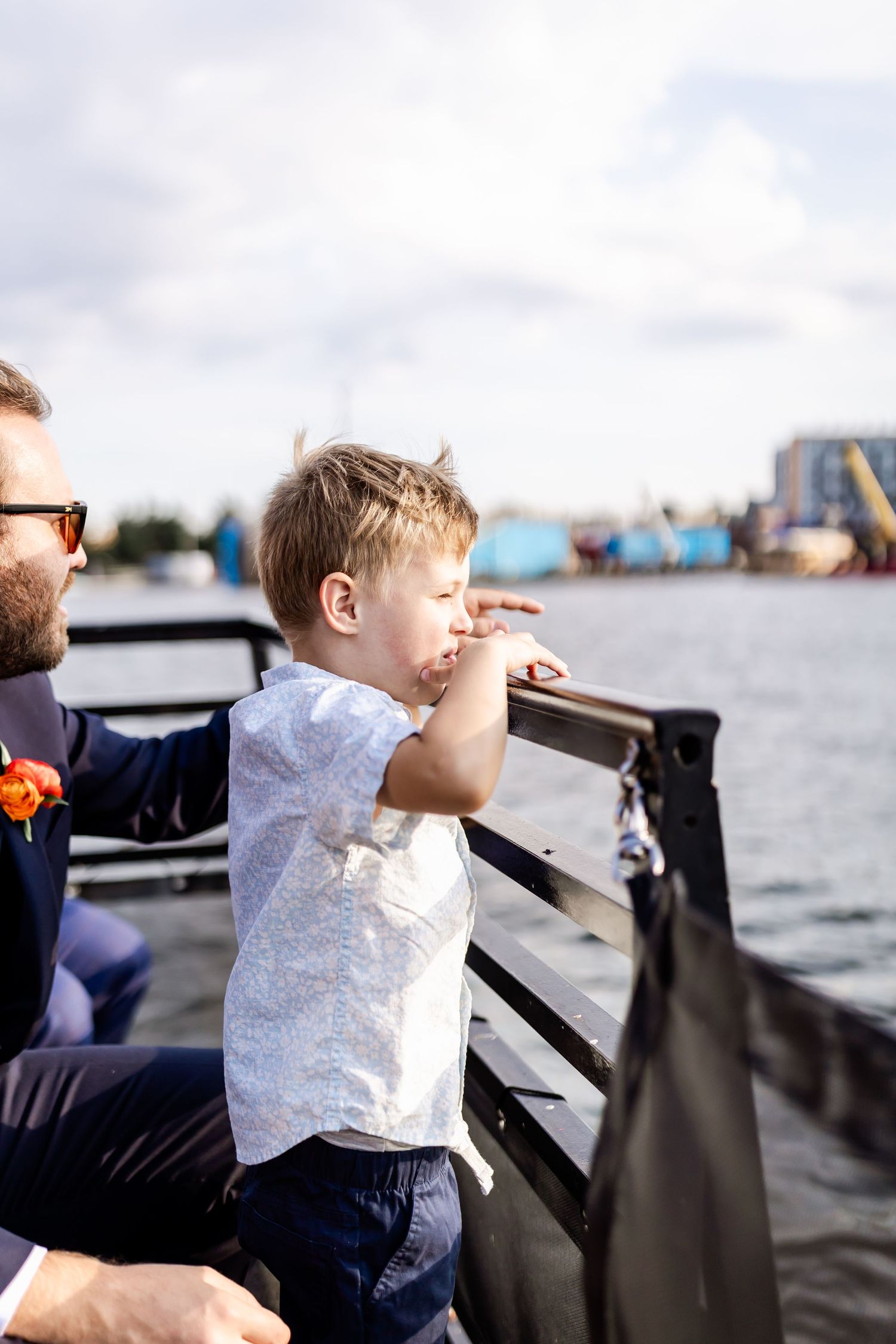 A child and adult stand at a waterfront railing overlooking an industrial harbor scene.