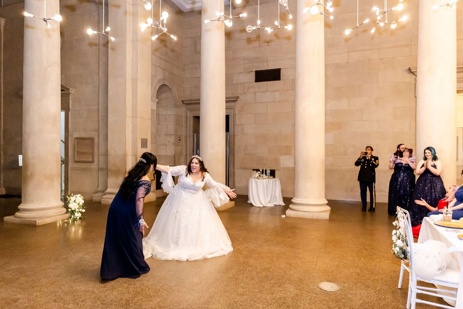 Wedding celebration in an elegant columned ballroom with hanging lights and seated guests.