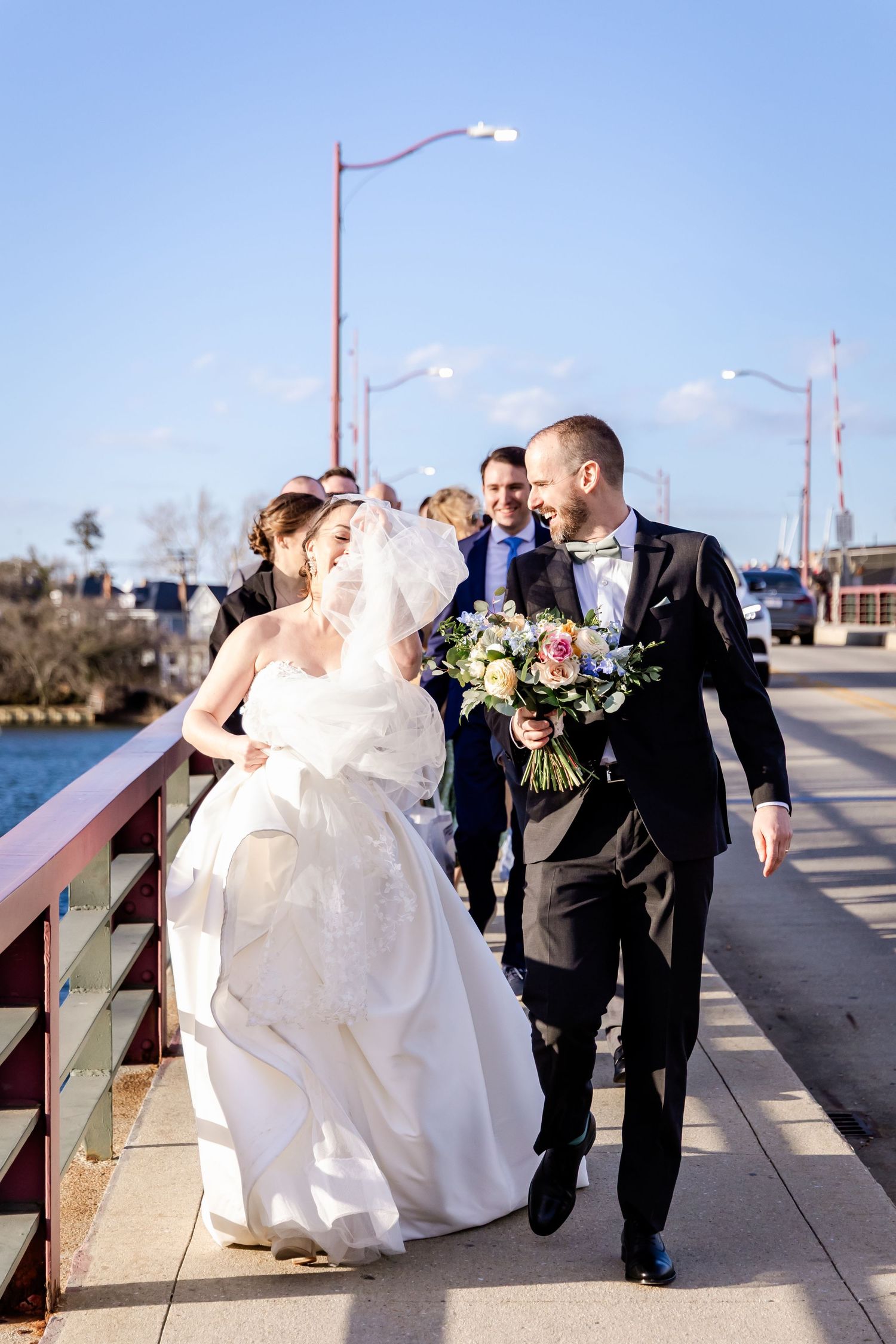 A wedding couple dances and spins on a wooden boardwalk by the water on a sunny day, their outfits flowing with movement.