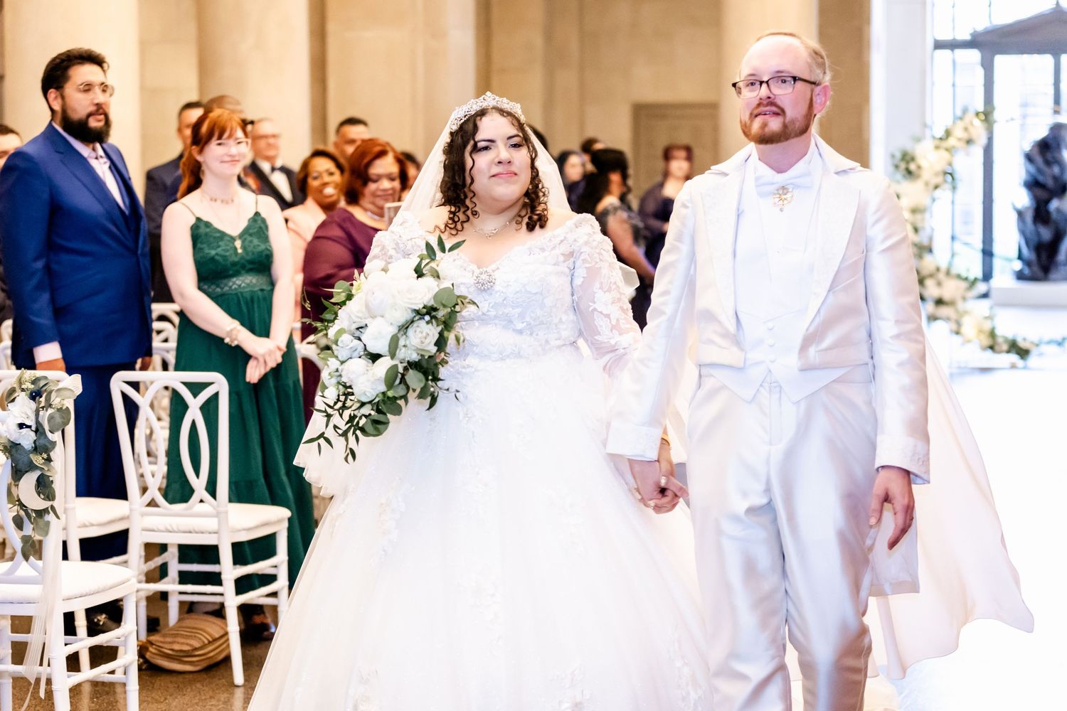 Wedding couple walking down the aisle in coordinated white outfits surrounded by seated guests.