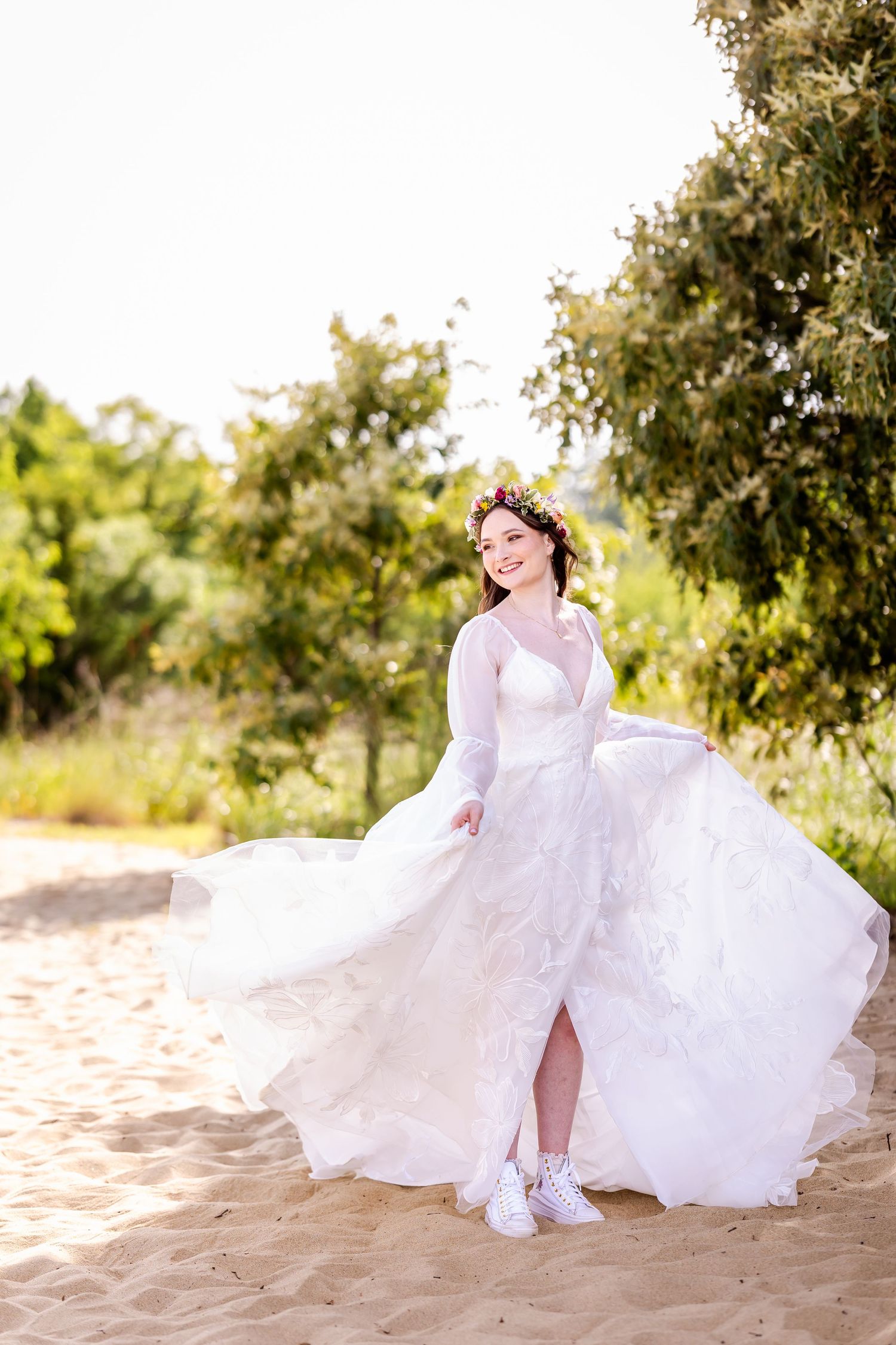 A bride in a flowing white dress twirls gracefully on a sandy beach during a sunset photo session.