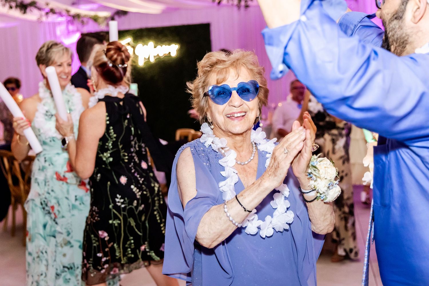 Wedding guests dance and celebrate on a festively lit dance floor with pink and purple lighting.