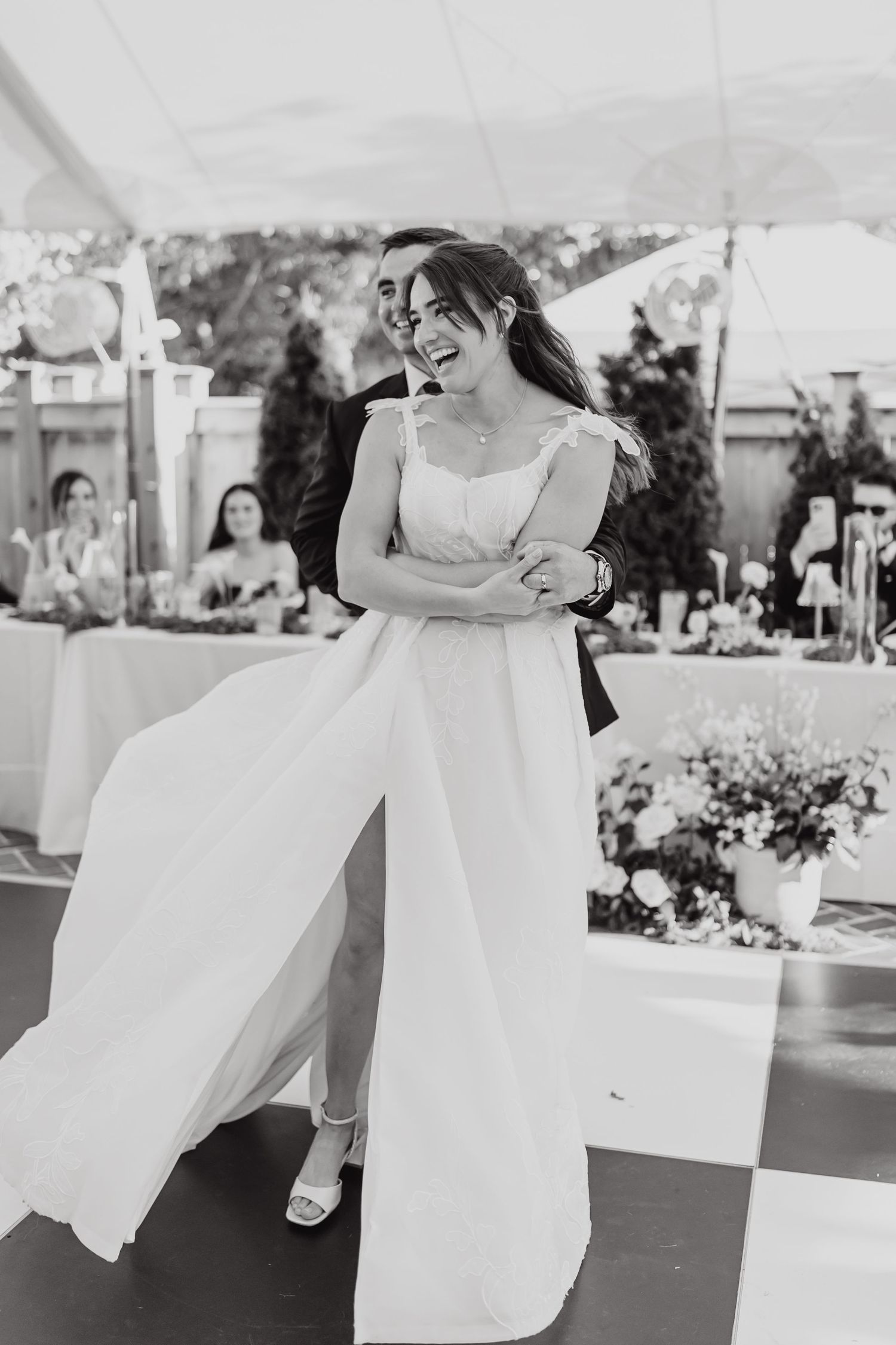 A bride in a flowing white jumpsuit laughs joyfully while dancing at an outdoor wedding reception.