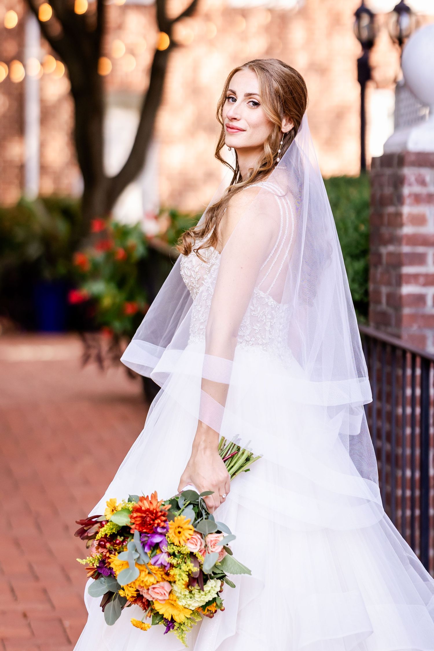 A bride in a white wedding dress holds a vibrant bouquet of yellow, orange and purple flowers on a brick walkway.