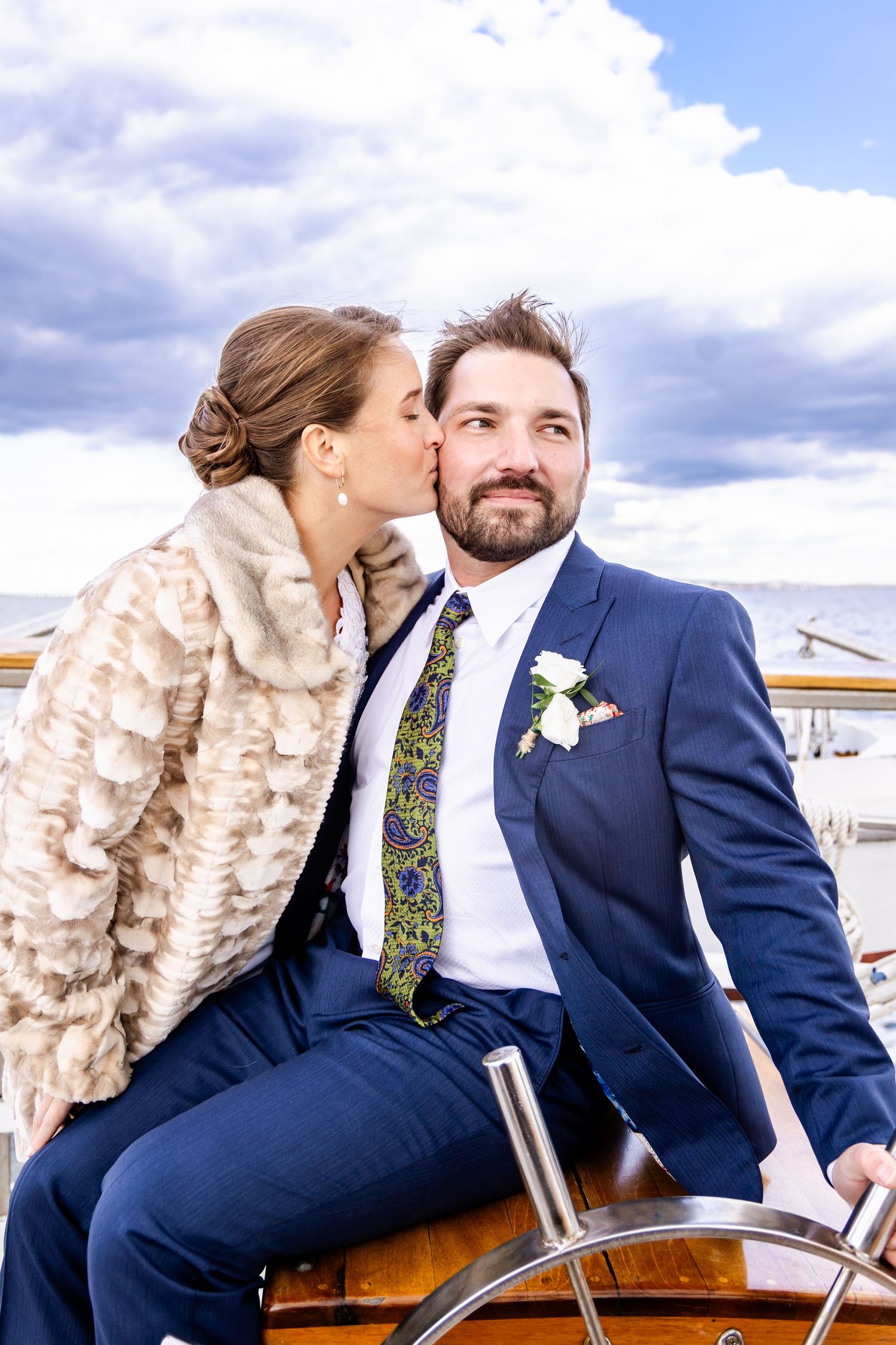 Bride kissing groom on cheek while he steers a wooden boat, wearing blue suit with boutonniere and patterned tie.